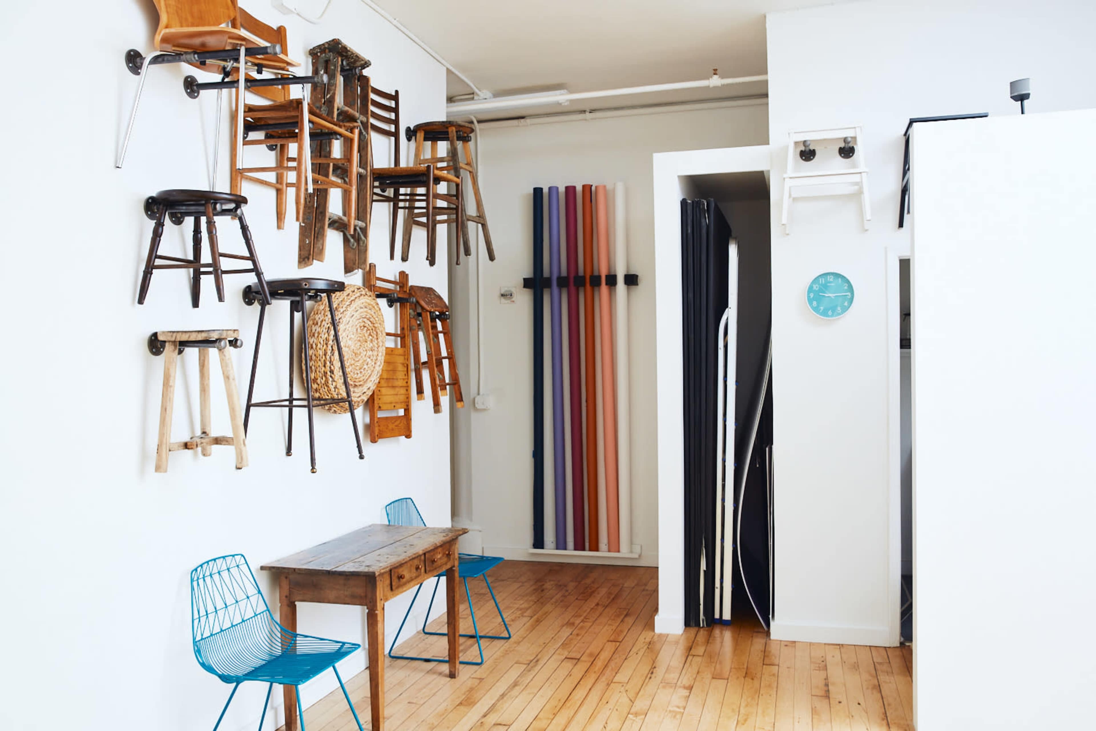 The image shows a bright room with a white wall displaying various wooden chairs mounted on it, along with a wooden table and blue wire chairs positioned on the floor.