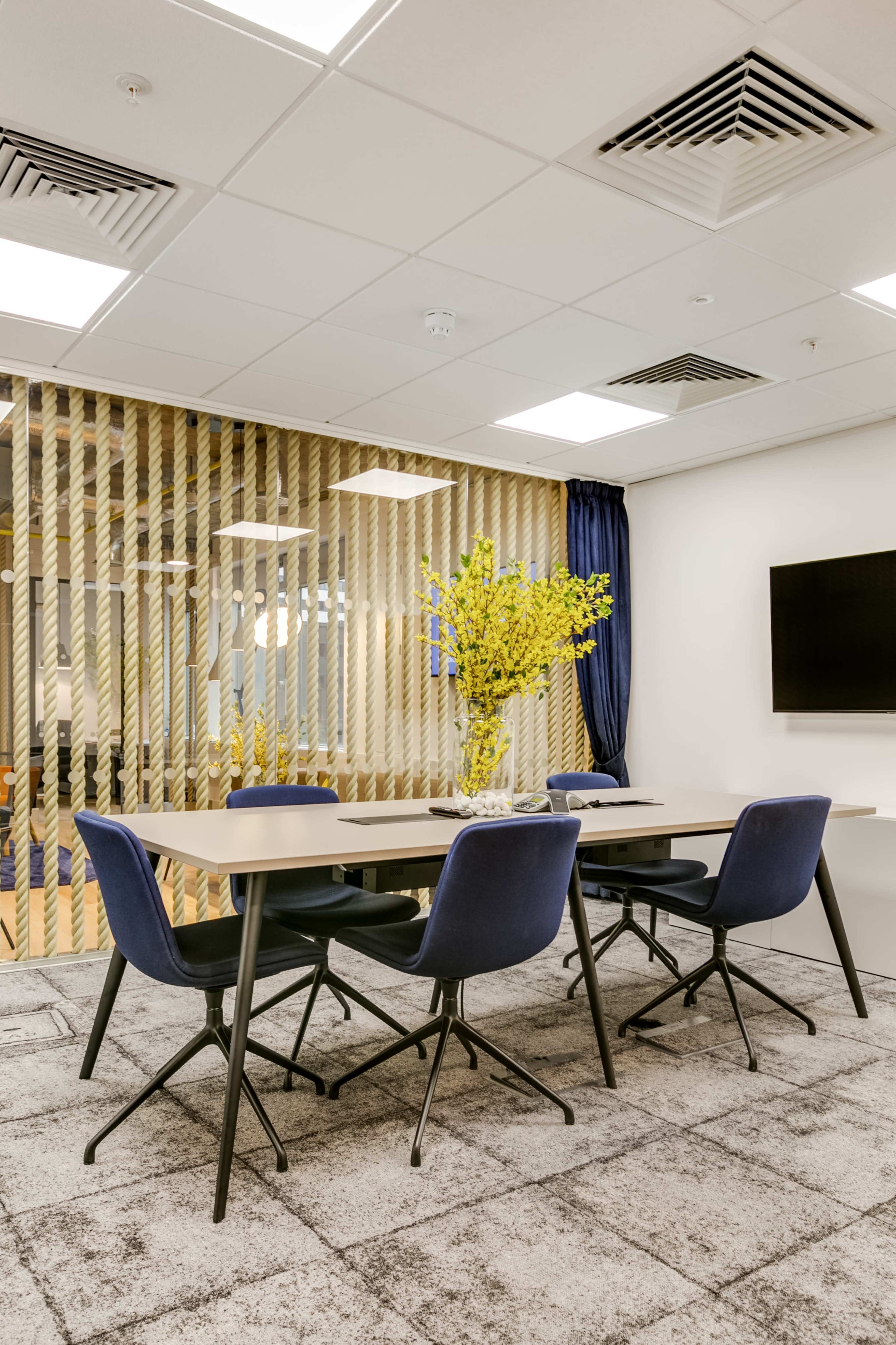 A modern meeting room features a rectangular table surrounded by four blue chairs, with a vase of yellow flowers as the centerpiece and a wall-mounted television in the background.