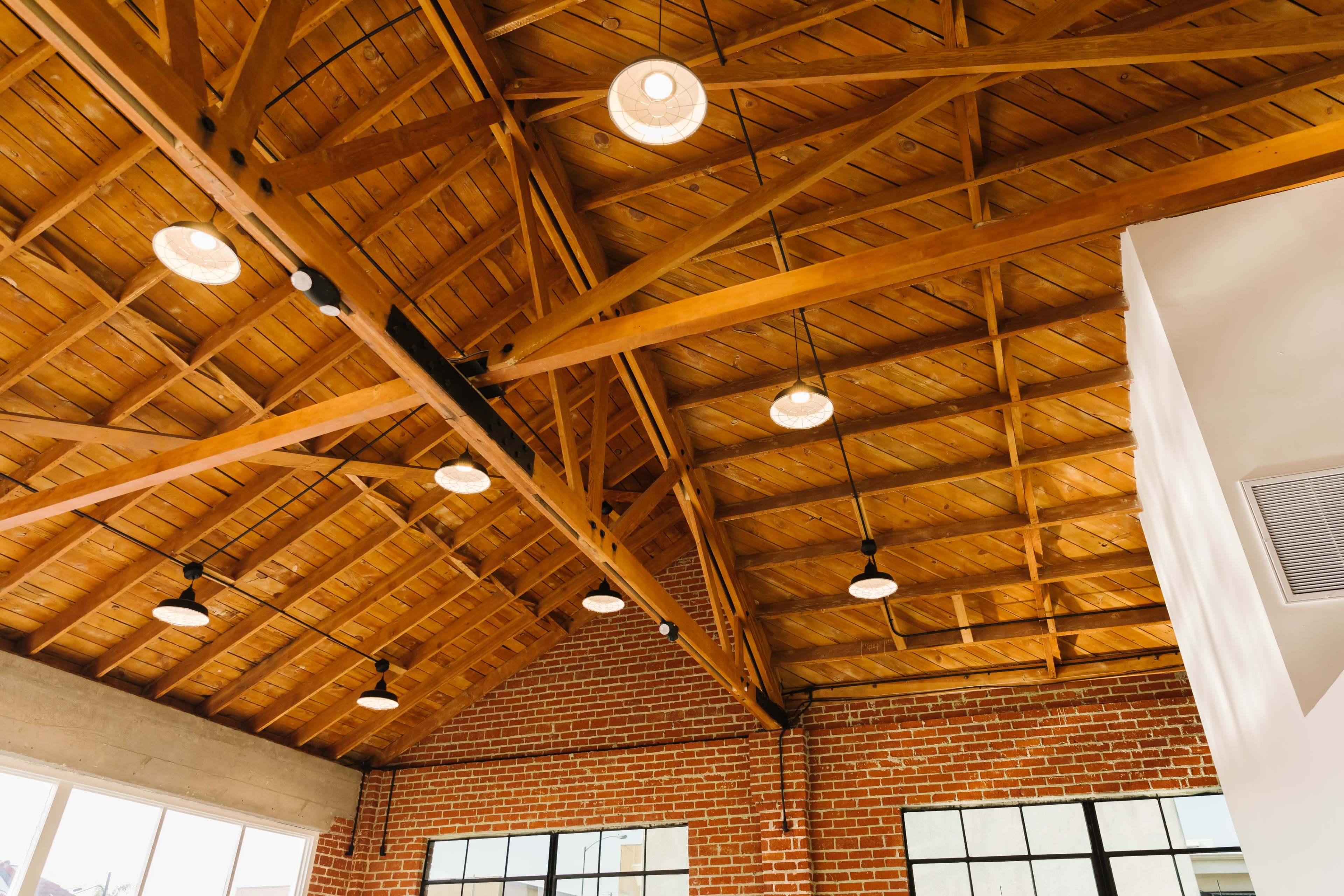 A spacious interior with a wooden beam ceiling and several circular pendant lights, accompanied by a brick wall and large windows.