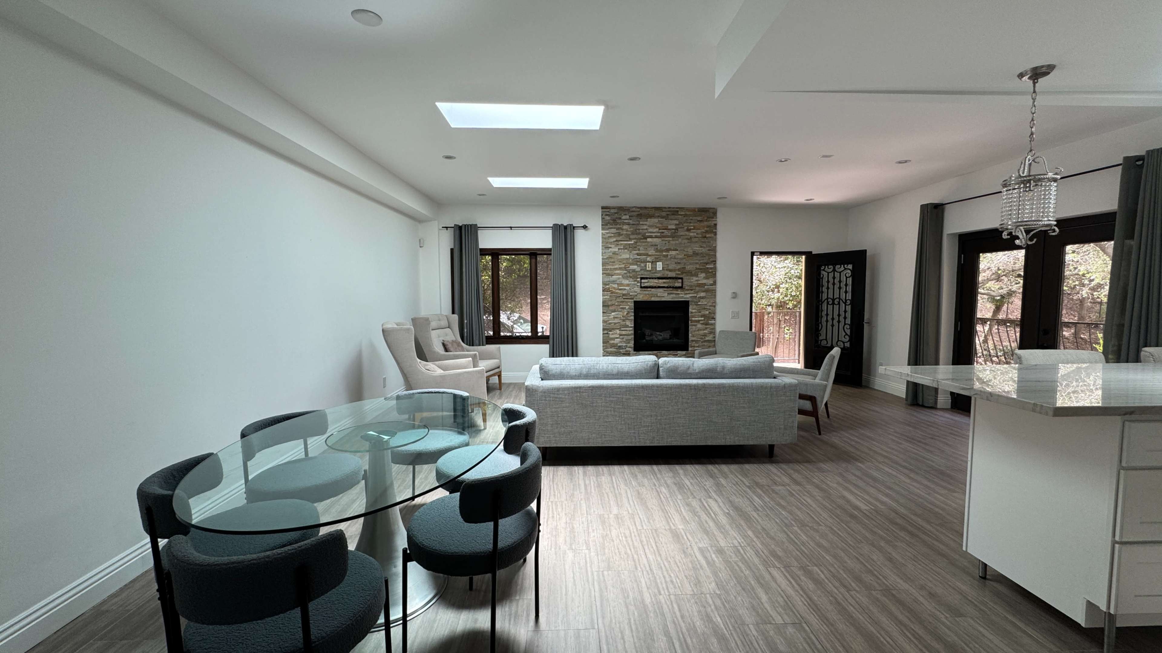 The image shows a modern living space featuring a glass dining table with black chairs, a comfortable gray sectional sofa, and a stone fireplace.