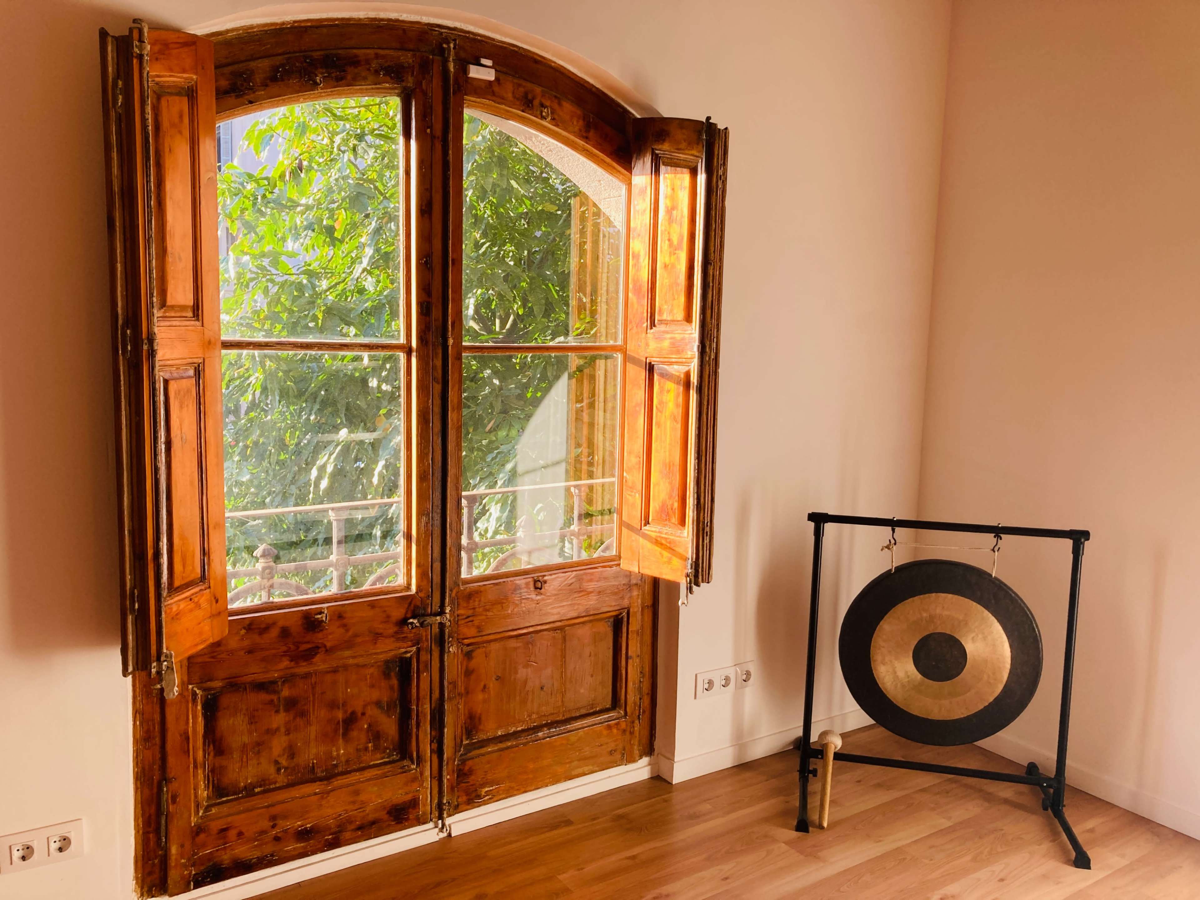 A pair of wooden double doors opens to reveal greenery outside, while a large gong stands on a black frame beside the doors in a well-lit room.