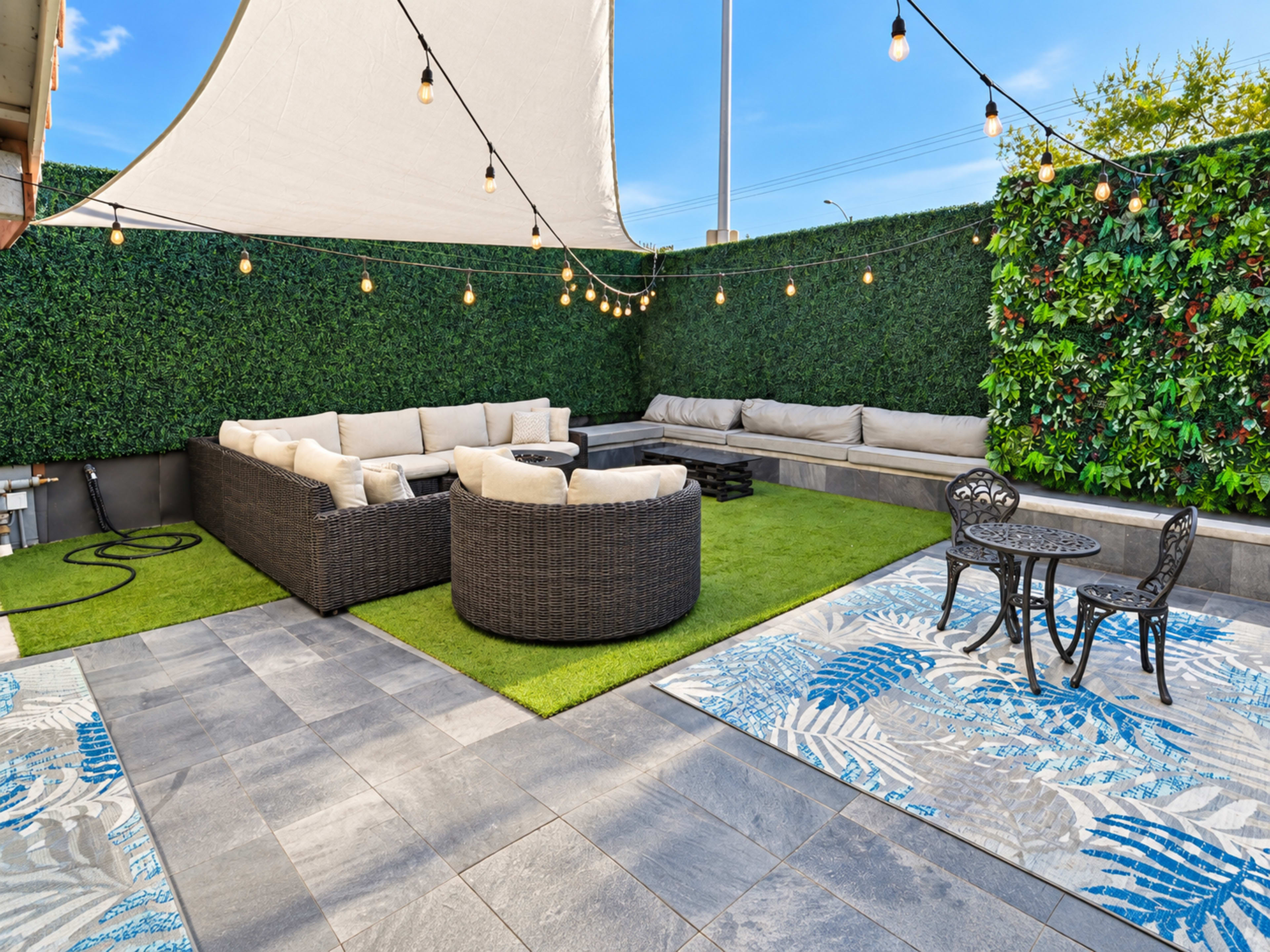 The image shows a landscaped outdoor patio area featuring a large seating arrangement with cushions, a grassy section, and a small table and chairs under string lights.