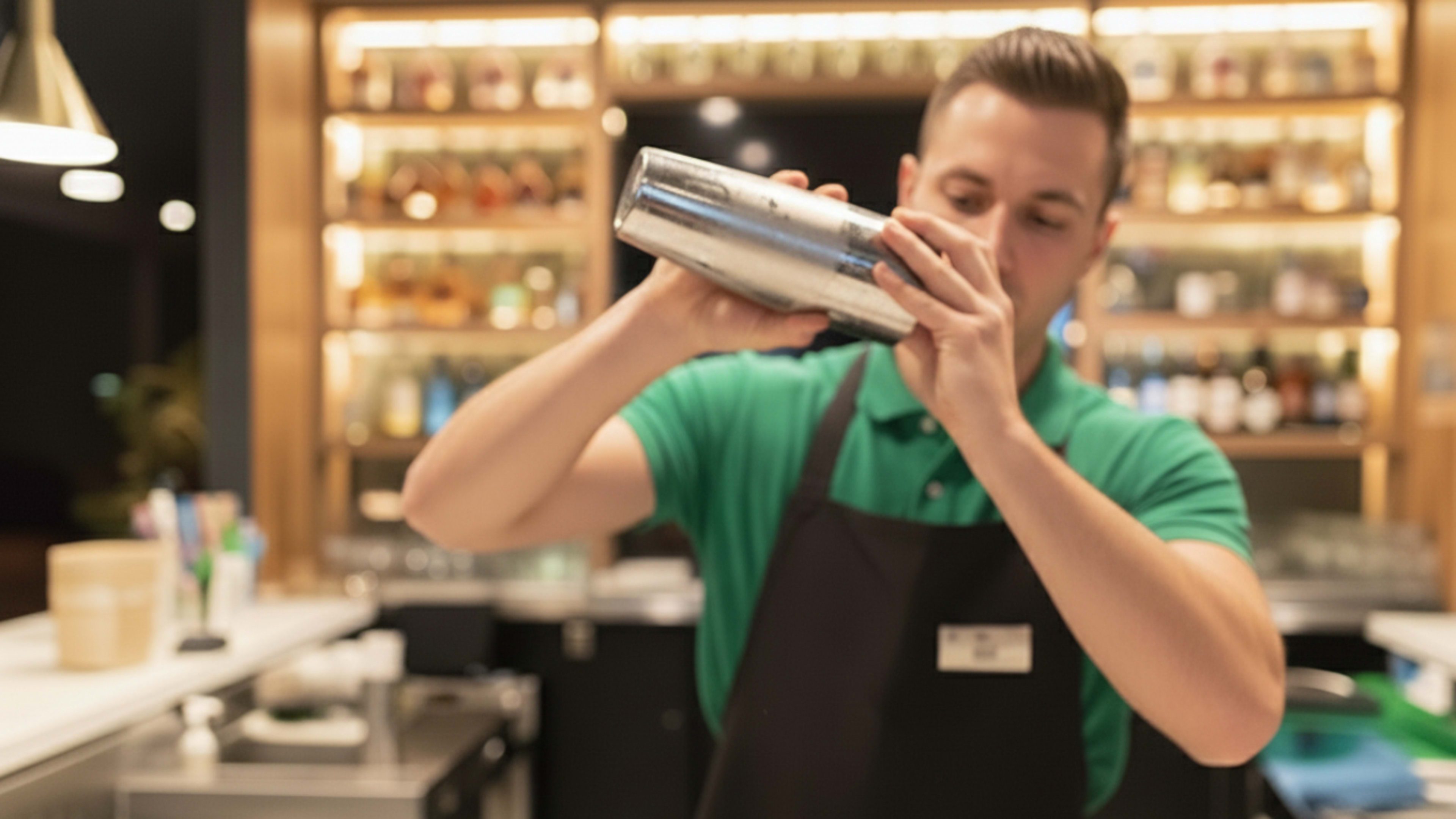 A bartender in a green shirt and black apron shakes a cocktail shaker behind a well-stocked bar.