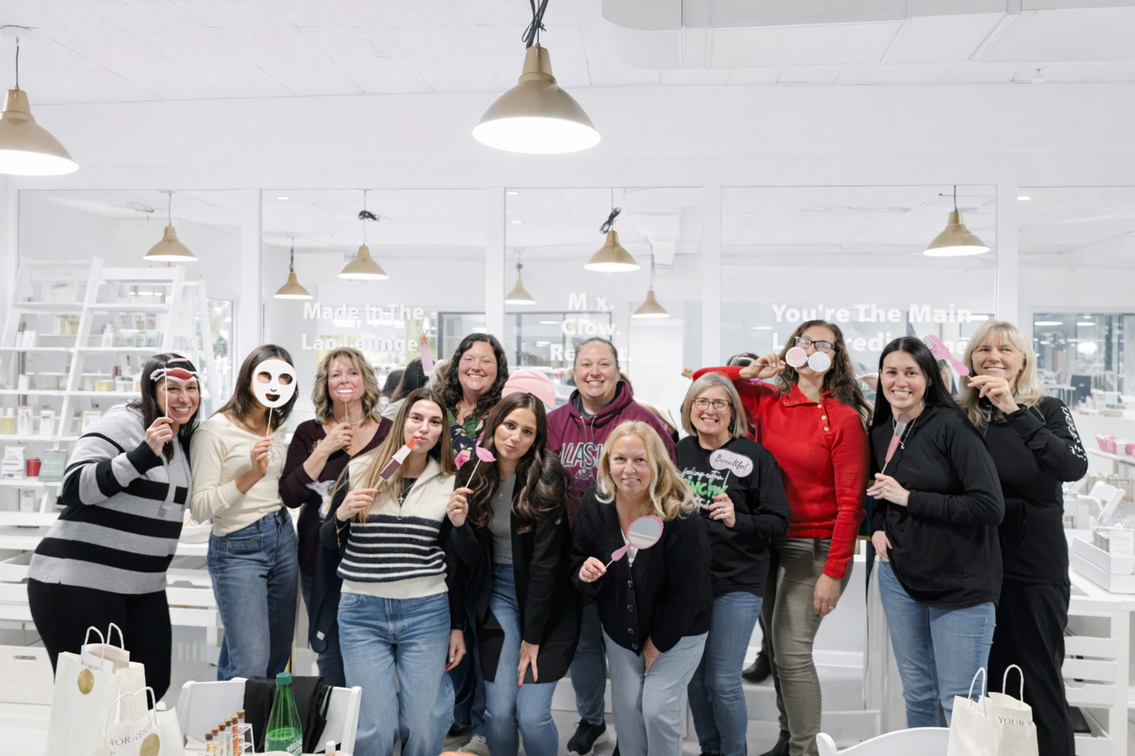 A group of twelve women stands together, smiling and posing with various props in a bright, modern indoor space.
