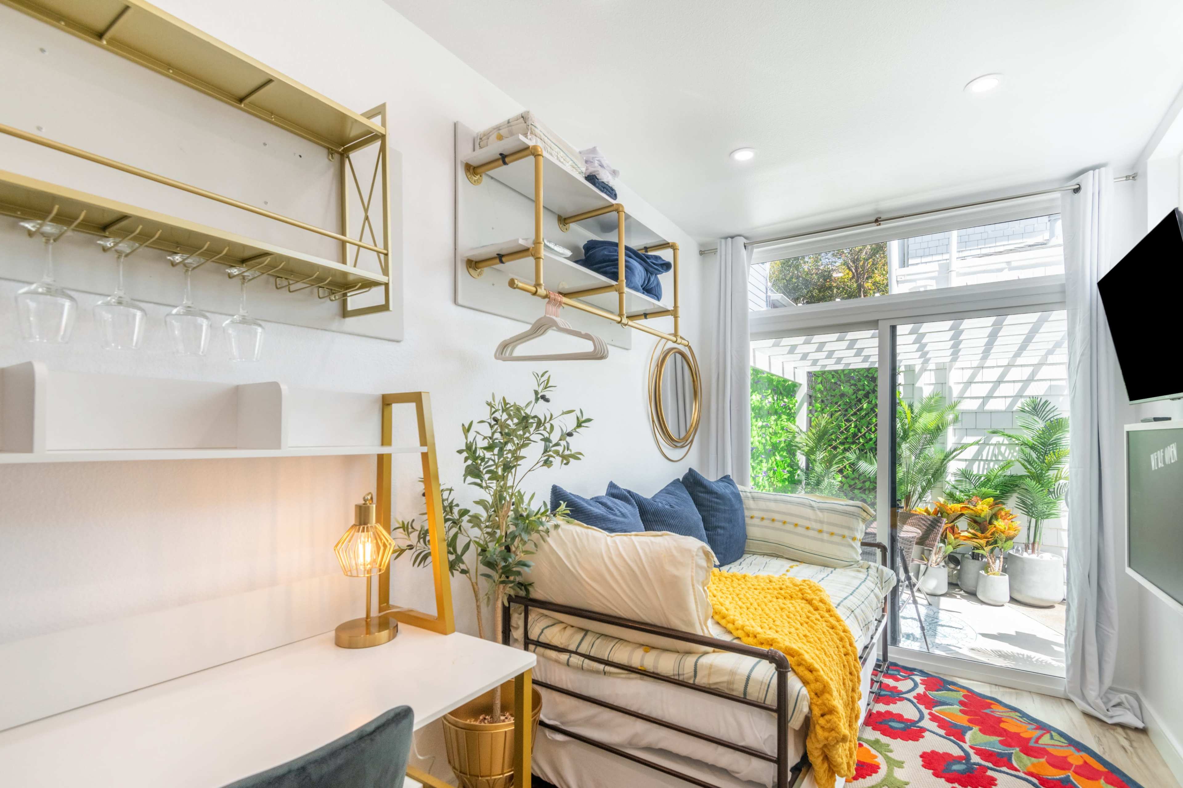 The image shows a bright living space featuring a sofa with colorful cushions, a desk with a lamp, and a sliding door leading to an outdoor area with plants.
