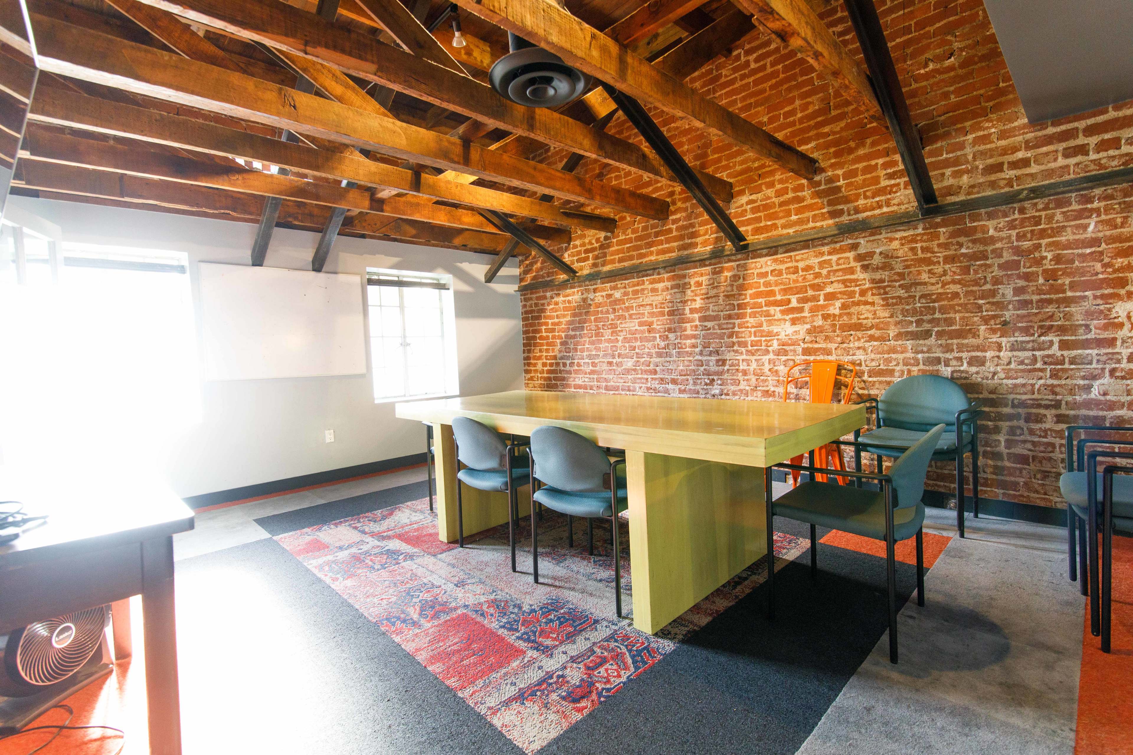 A modern conference room featuring a large wooden table surrounded by chairs, with exposed wooden beams and brick walls.
