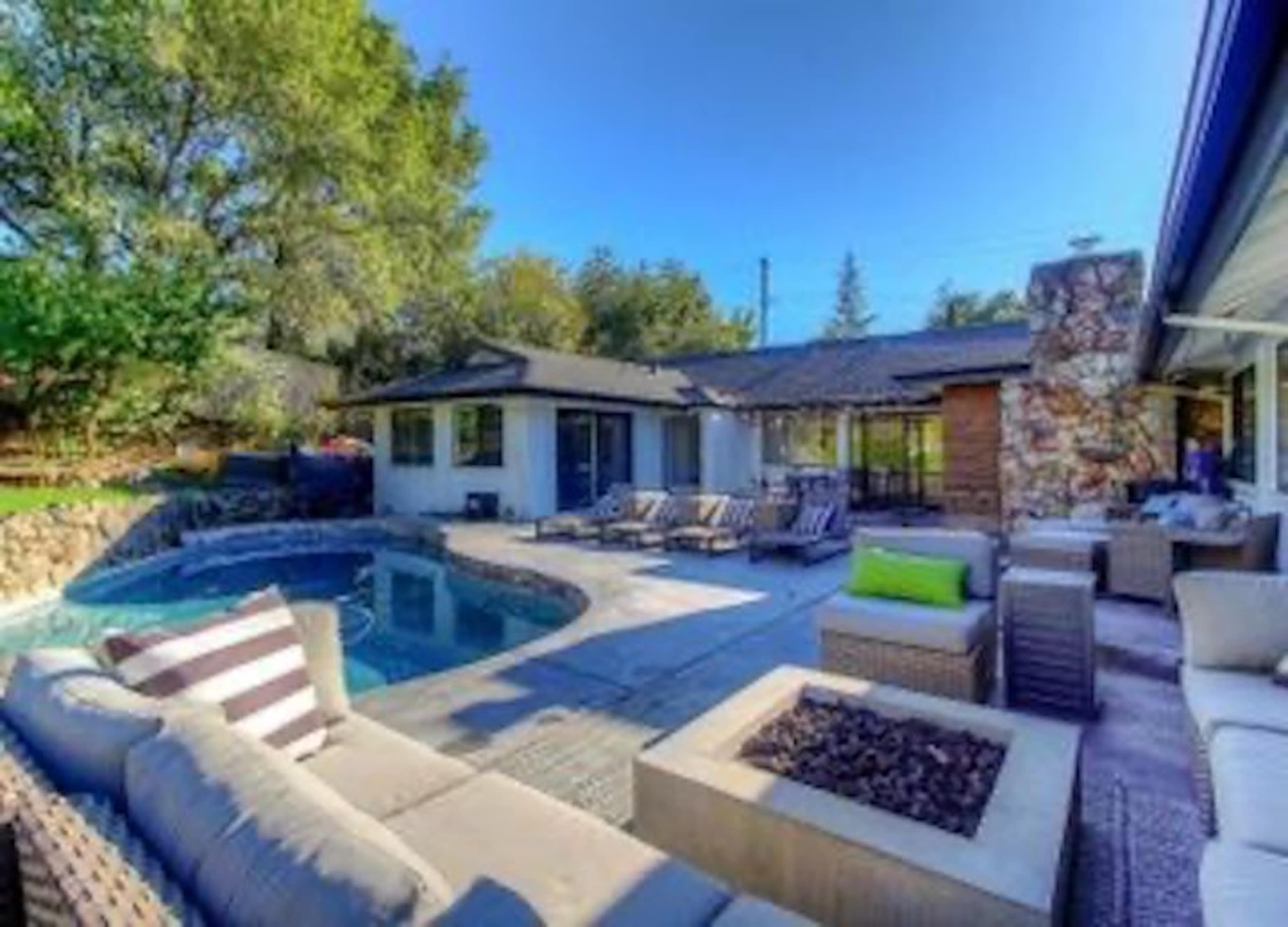 A spacious backyard features a swimming pool surrounded by lounge chairs and a stone patio, with a modern home and landscaped greenery in the background.