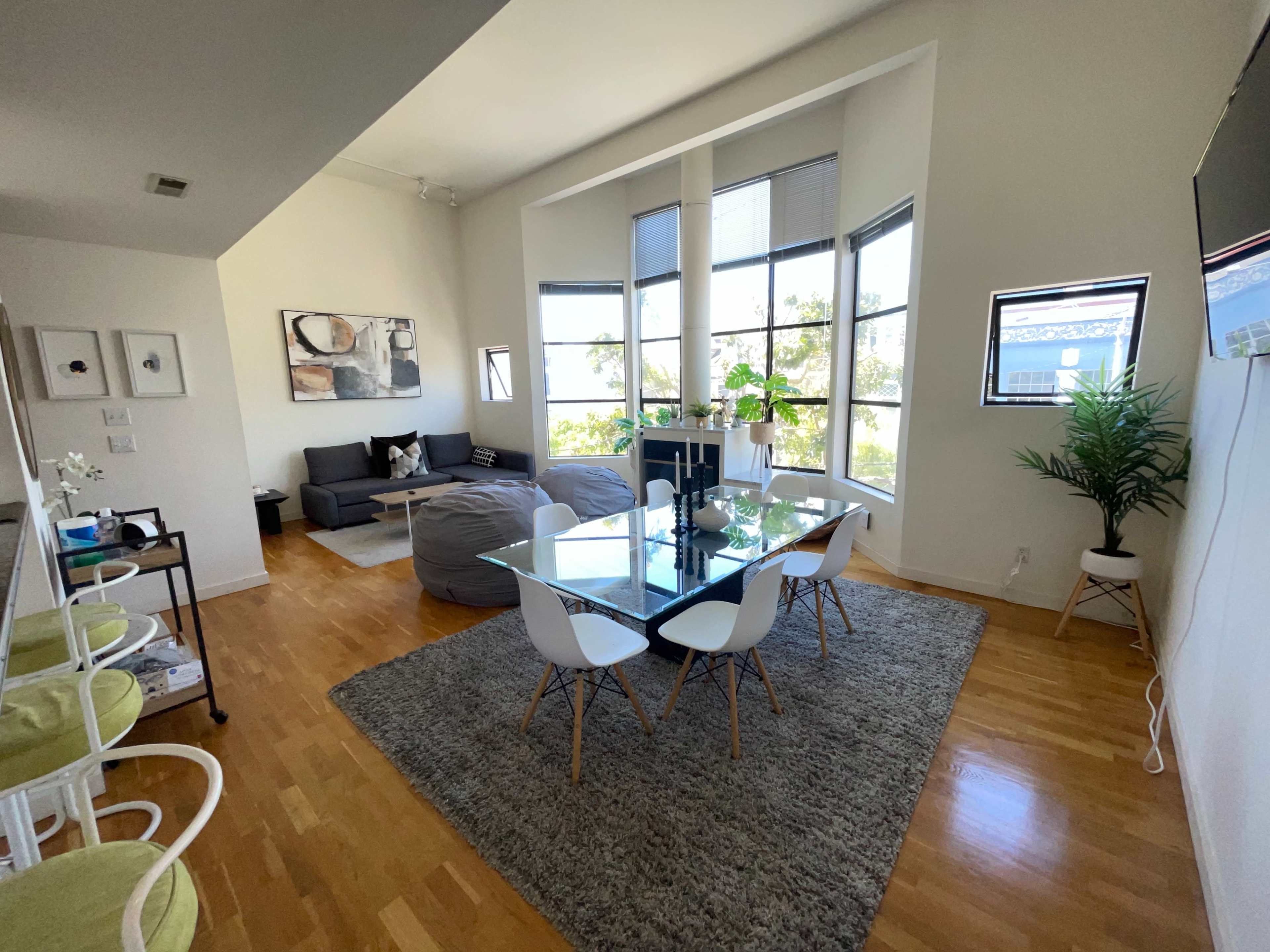 The image depicts a modern living space featuring a dining area with a glass table and white chairs, a gray sectional couch, and large windows allowing natural light.