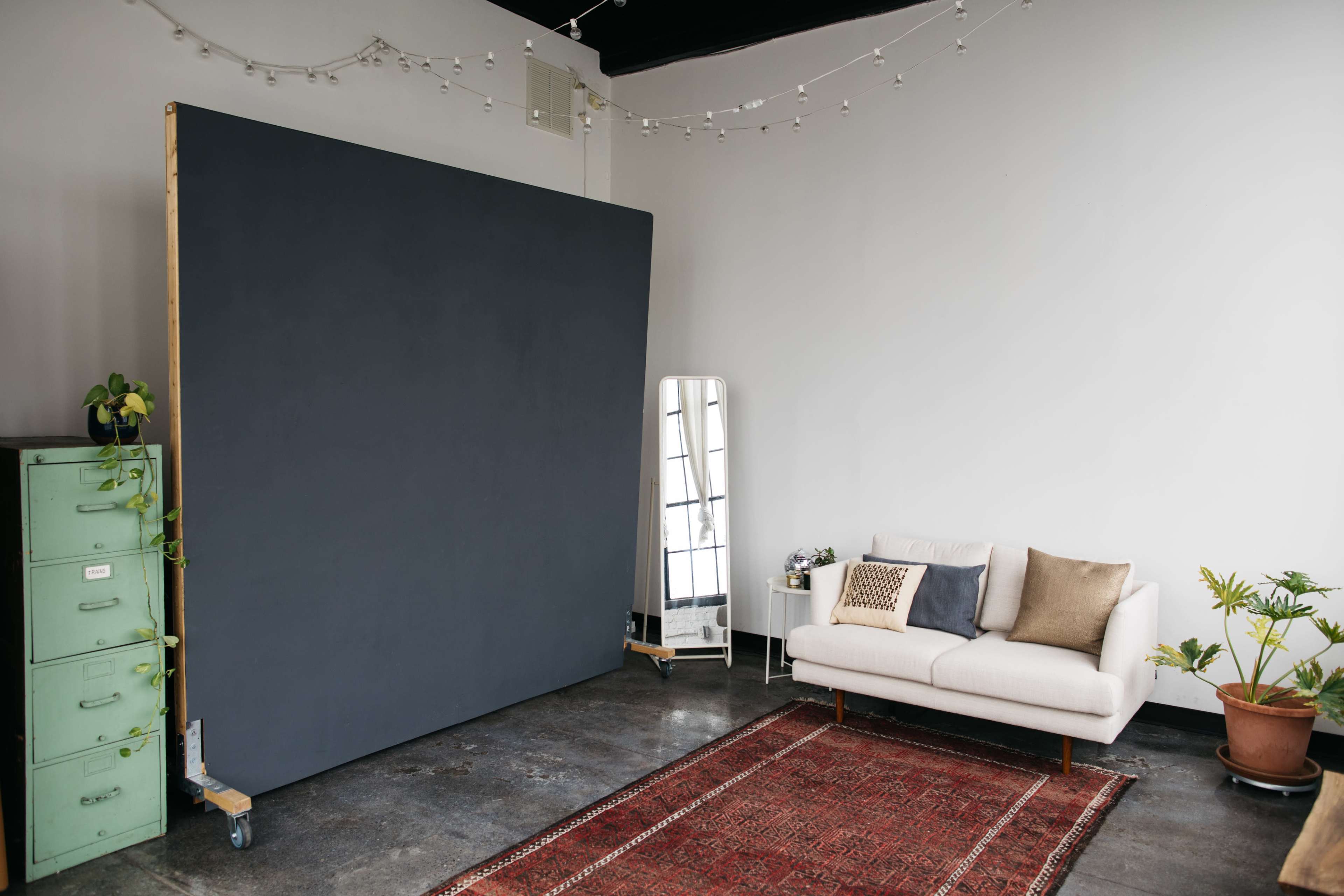 The image shows a minimalistic room with a large dark backdrop, a white couch with decorative pillows, a full-length mirror, and plants, all set on a patterned rug.