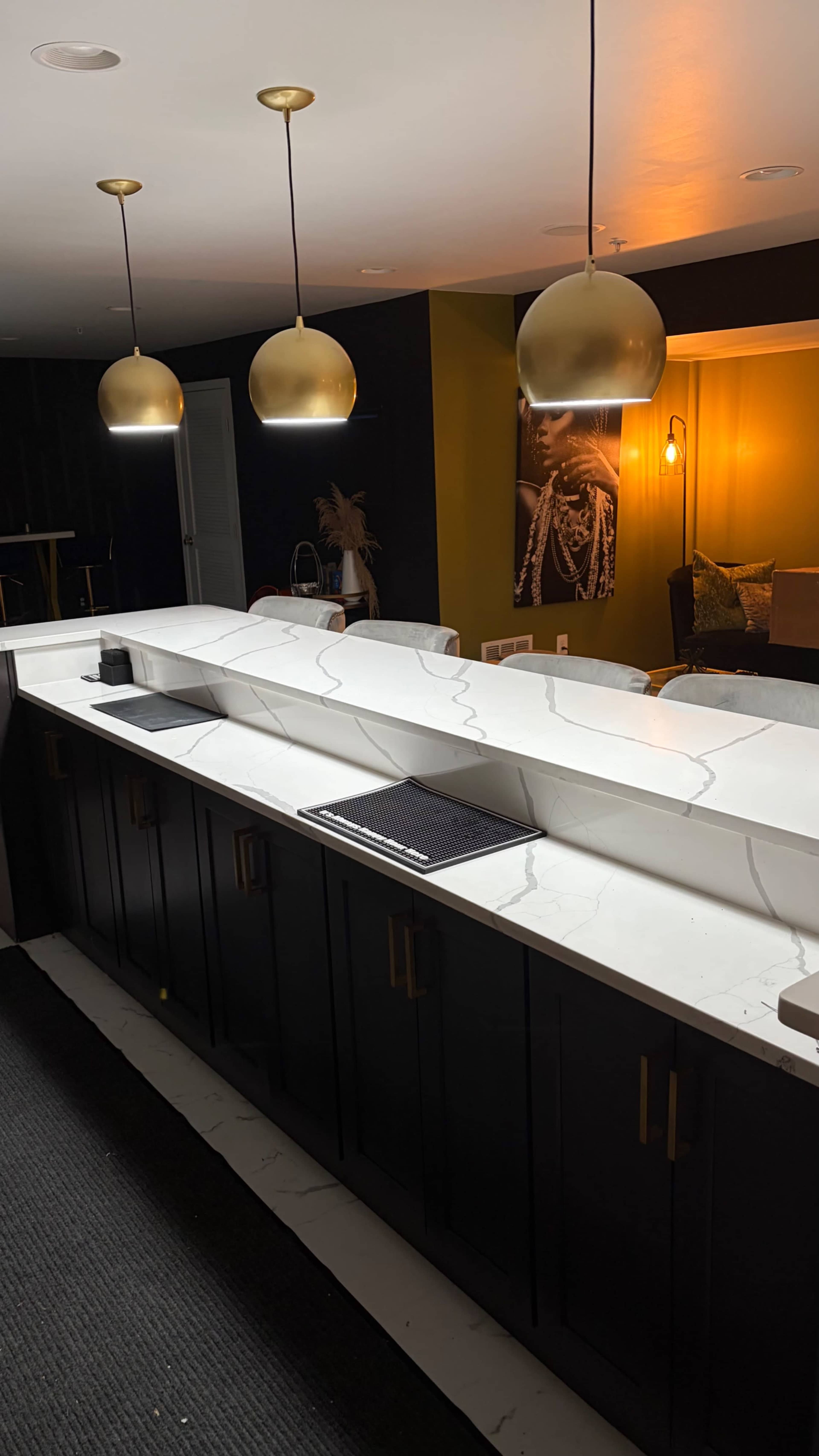 The image shows a modern kitchen with a large, white countertop and three pendant lights above, featuring dark cabinets and an open living room area in the background.