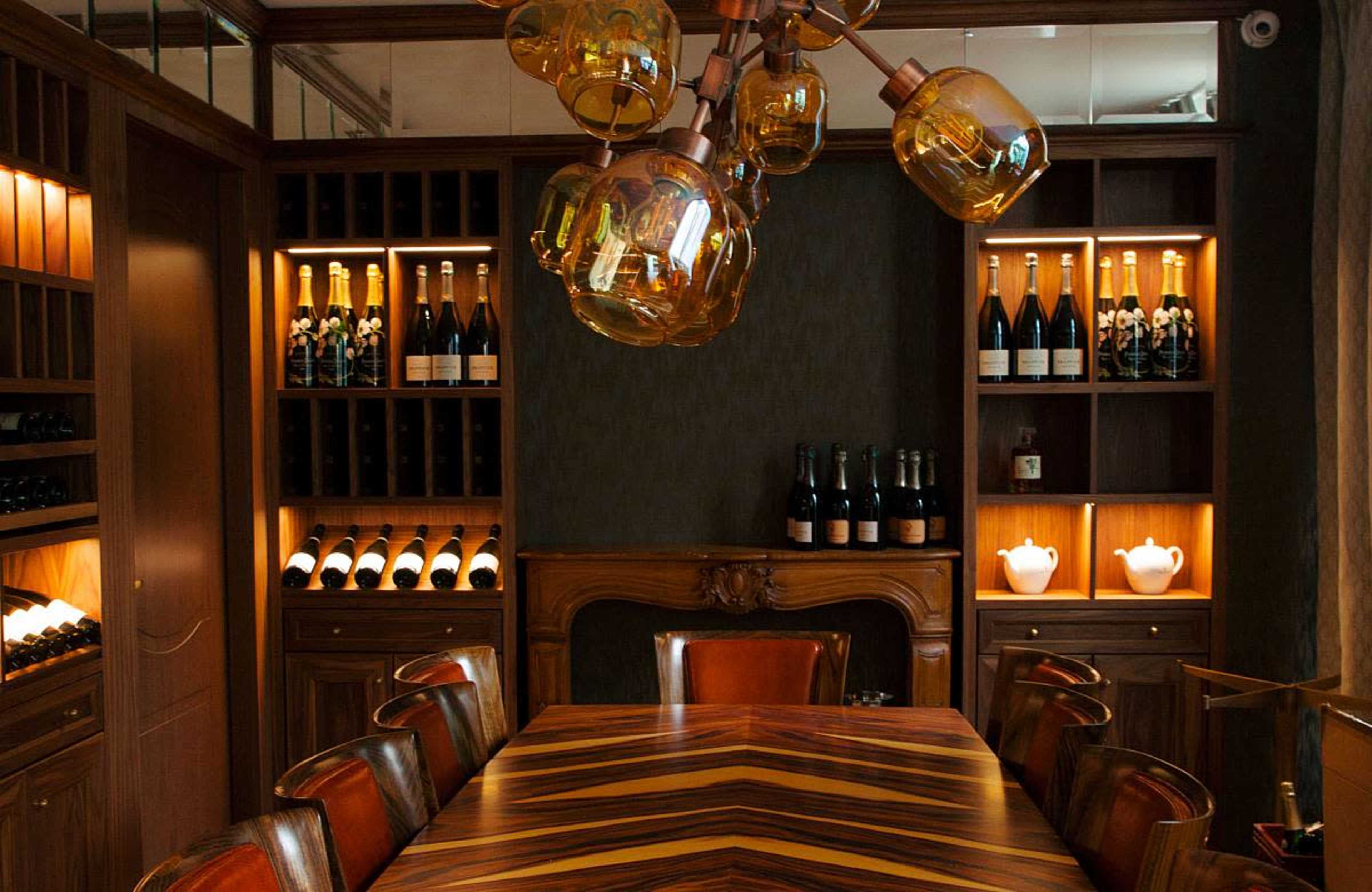 The image shows a private dining room featuring a wooden table surrounded by leather chairs, with shelves of champagne bottles and decorative lighting.