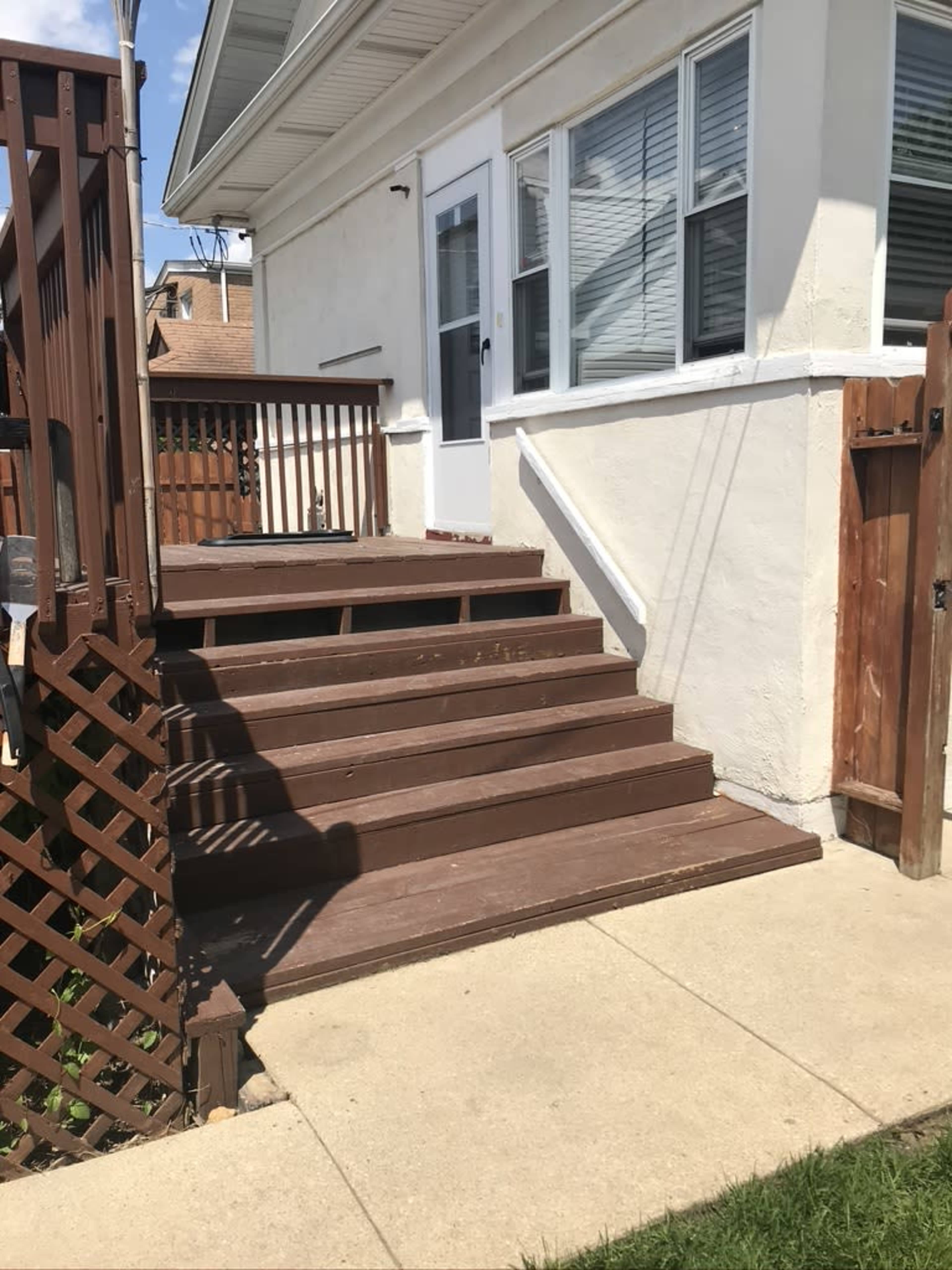 The image shows a back entrance to a house with wooden steps leading up to a door, surrounded by a railing and a lattice fence.