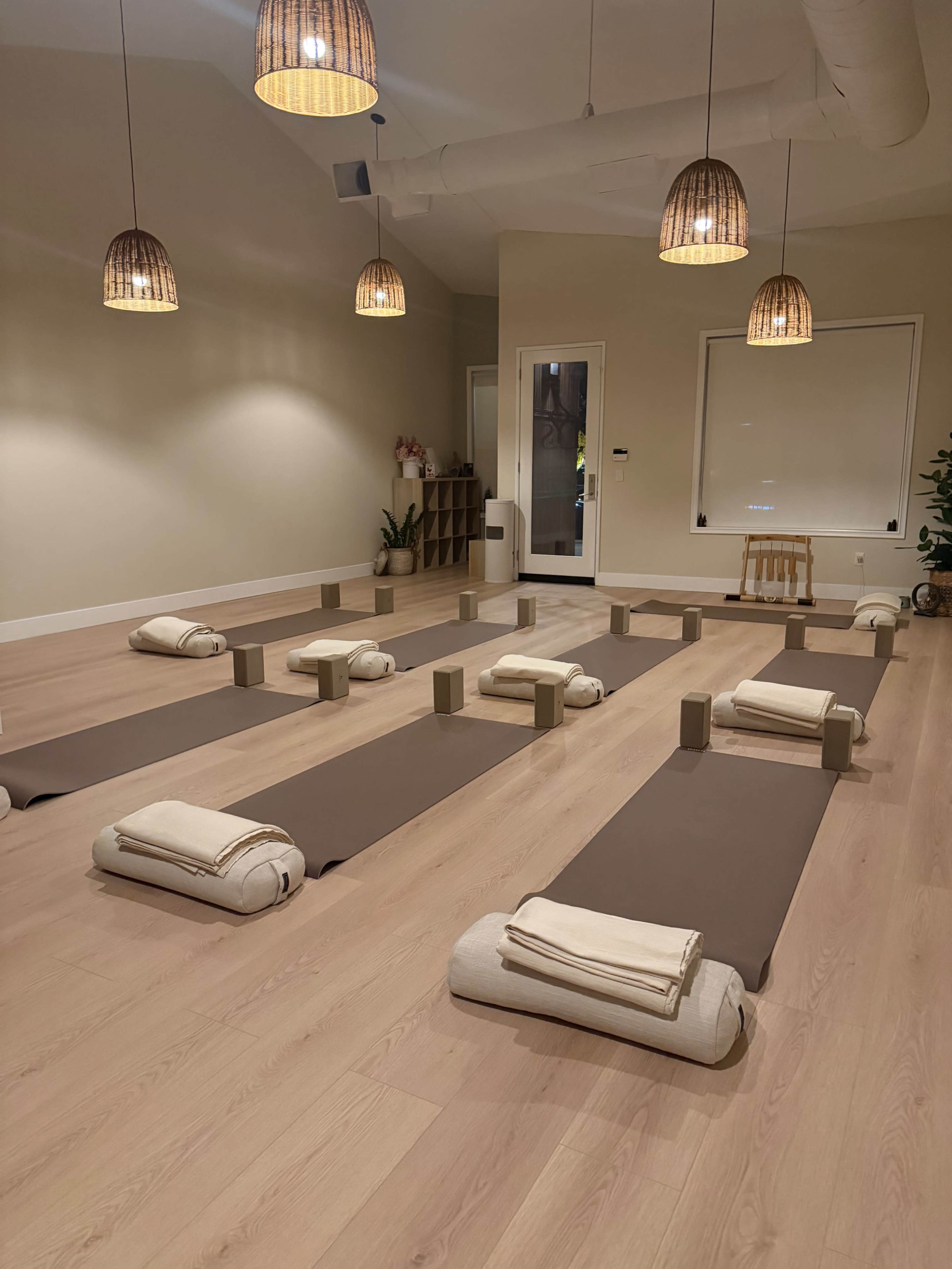 The image shows a serene yoga studio with neatly arranged mats and bolsters, ambient lighting from pendant lamps, and a calming neutral color scheme.