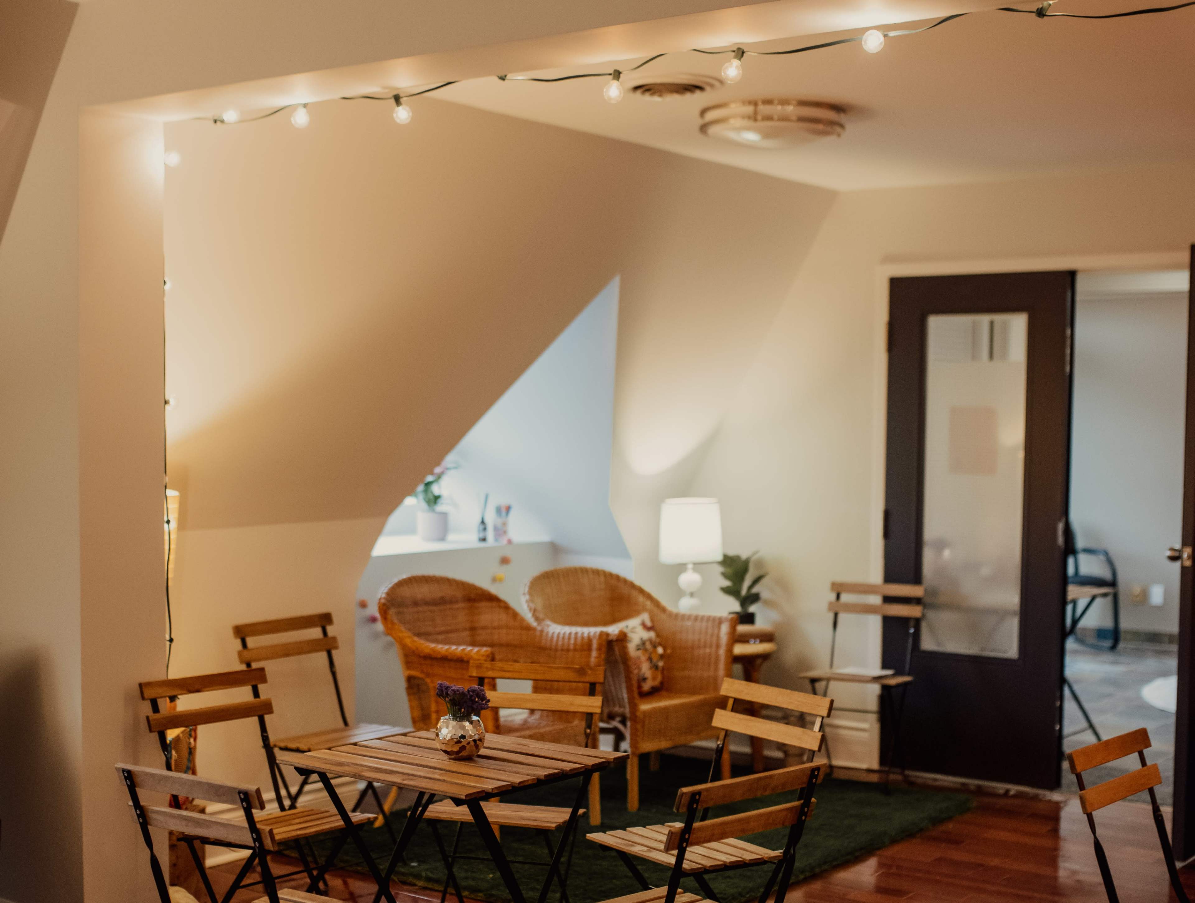 A cozy room features wooden tables and chairs arranged around a small seating area with wicker chairs and a lamp.