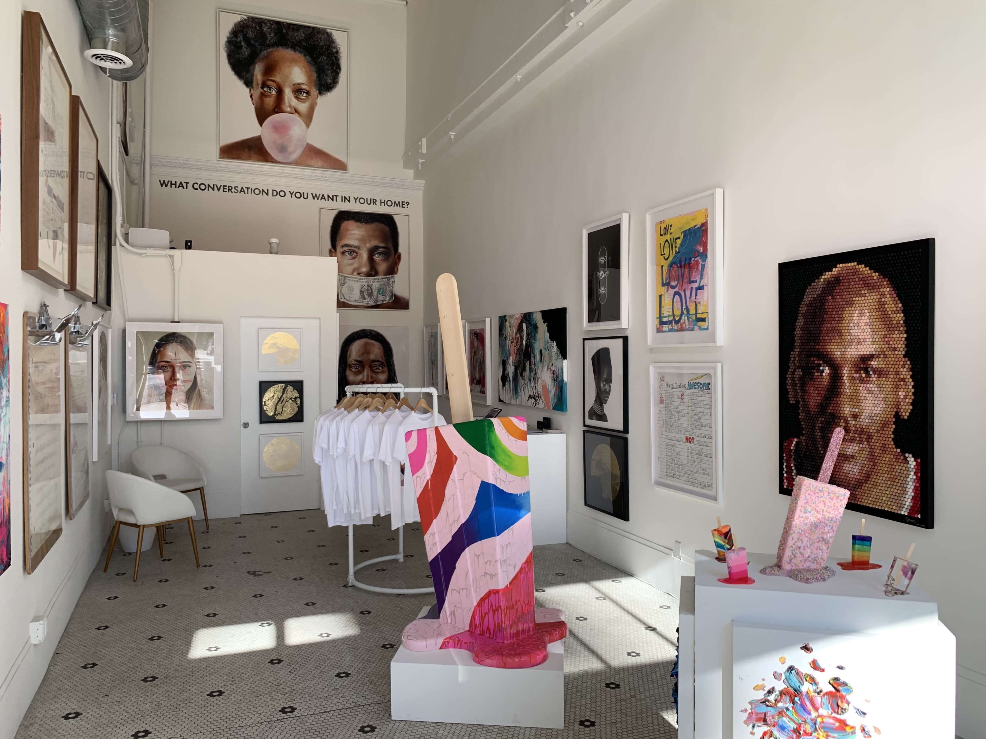 An art gallery featuring various artworks on the walls, including portraits and colorful installations, along with a display of white T-shirts on a rack.