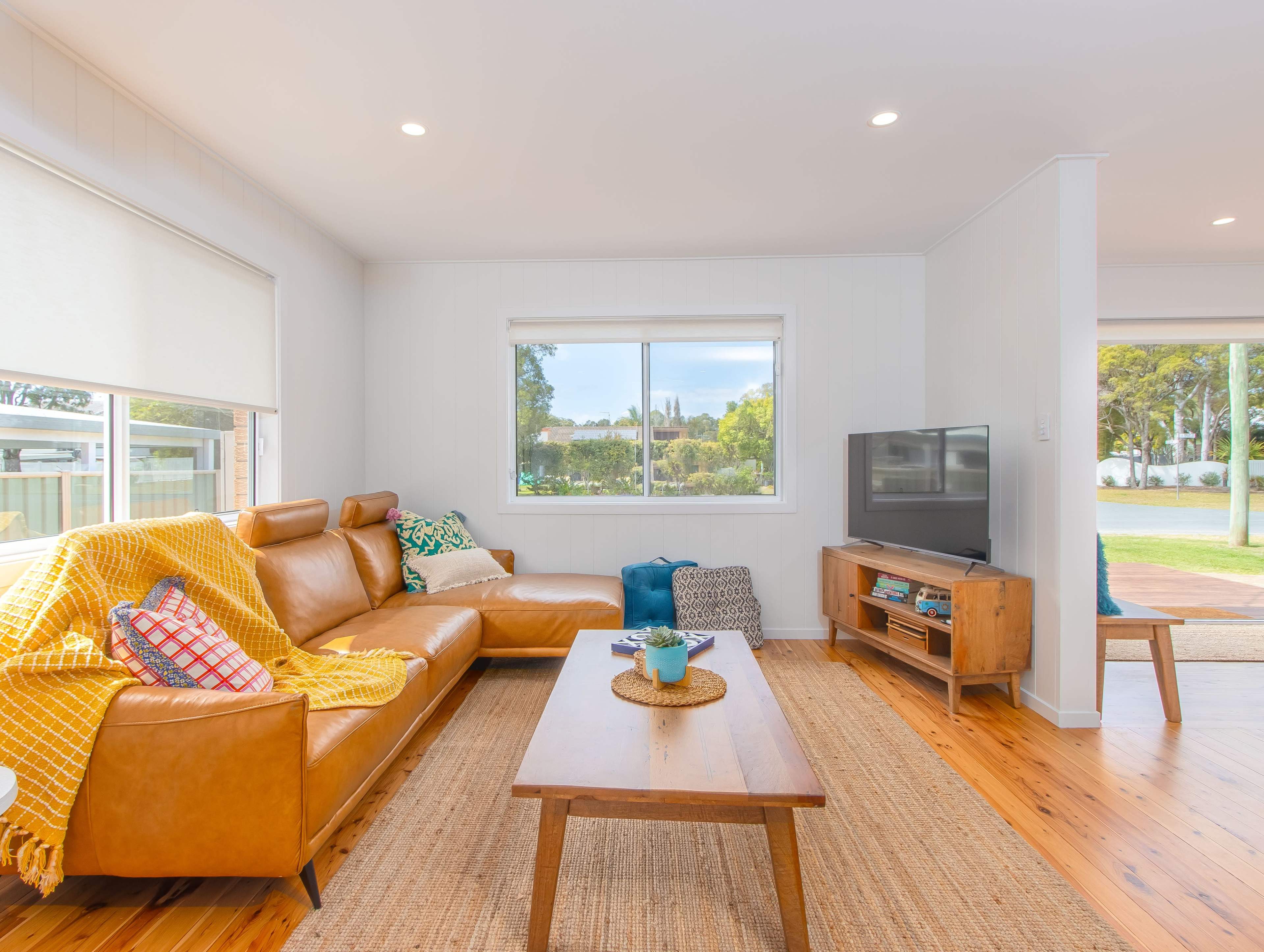 A cozy living room features a brown leather sofa, a wooden coffee table, and a television, with large windows providing a view of the outdoors.