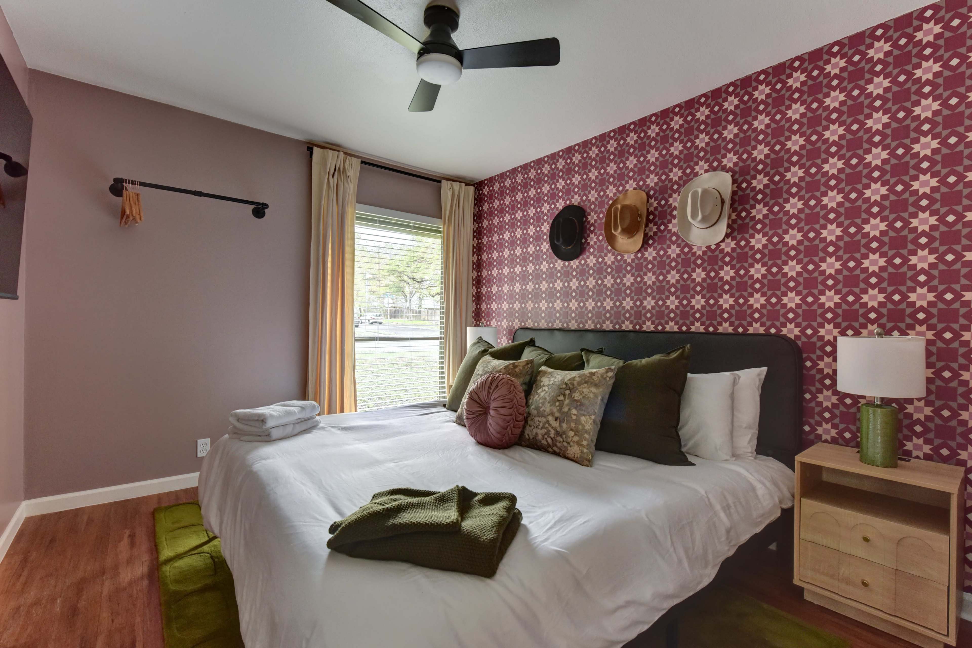 The image shows a neatly arranged bedroom with a large bed, decorative pillows, a nightstand, and patterned wallpaper featuring mounted hats.