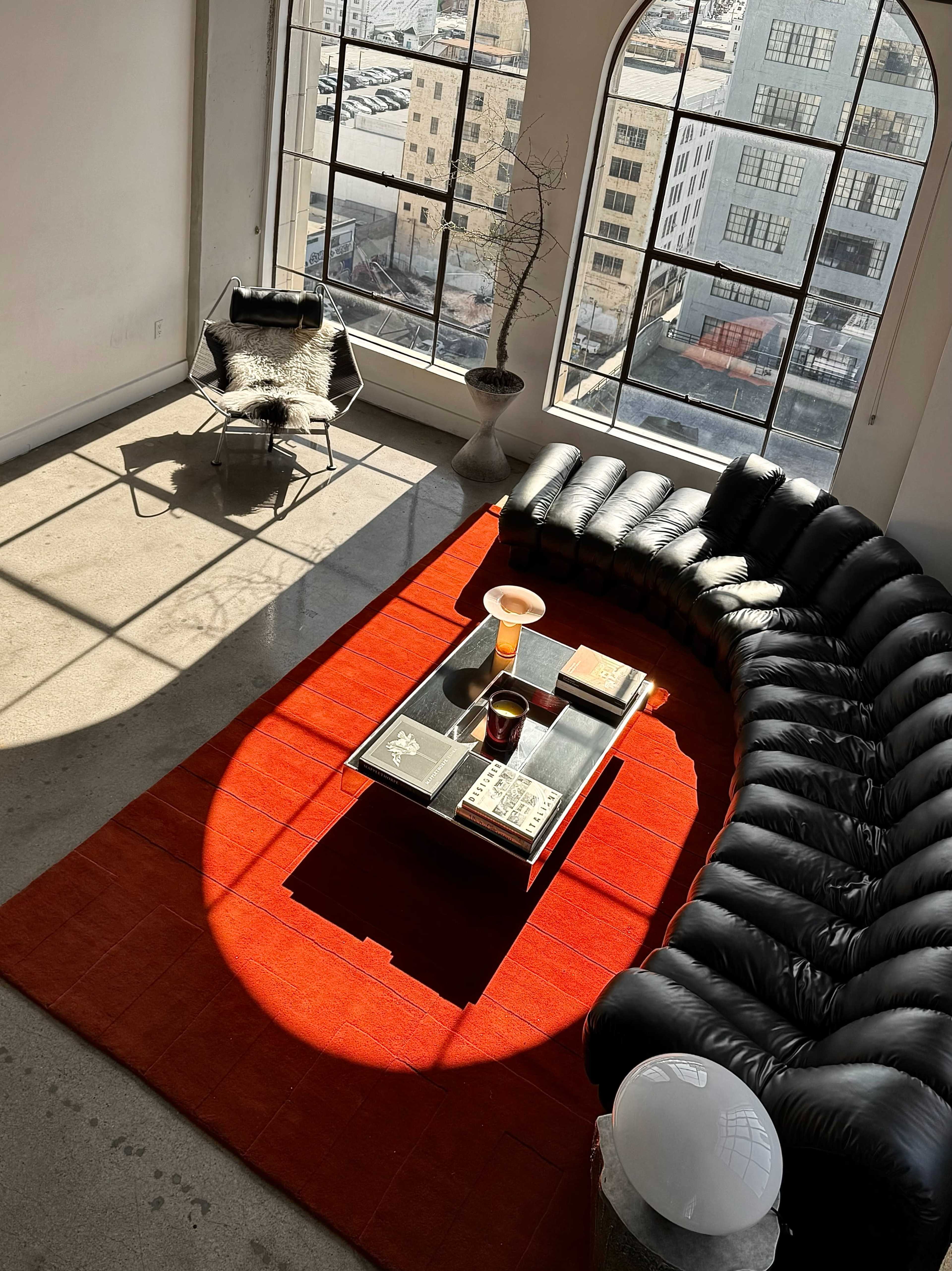 A curved black sofa surrounds a low table on a bright red rug in a well-lit room with large windows.