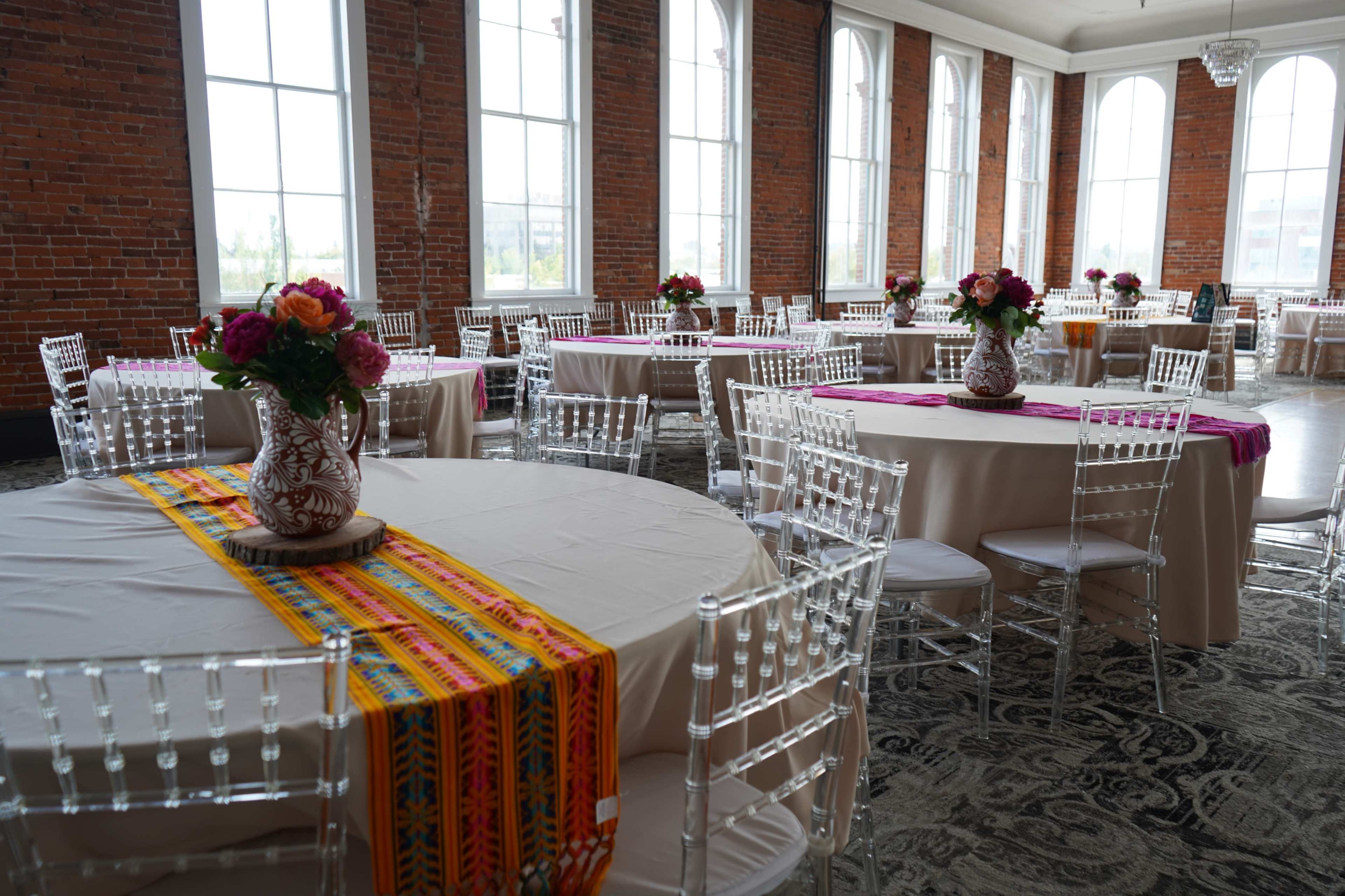 The image shows a banquet hall with round tables adorned with floral centerpieces and colorful table runners against a backdrop of large windows and exposed brick walls.