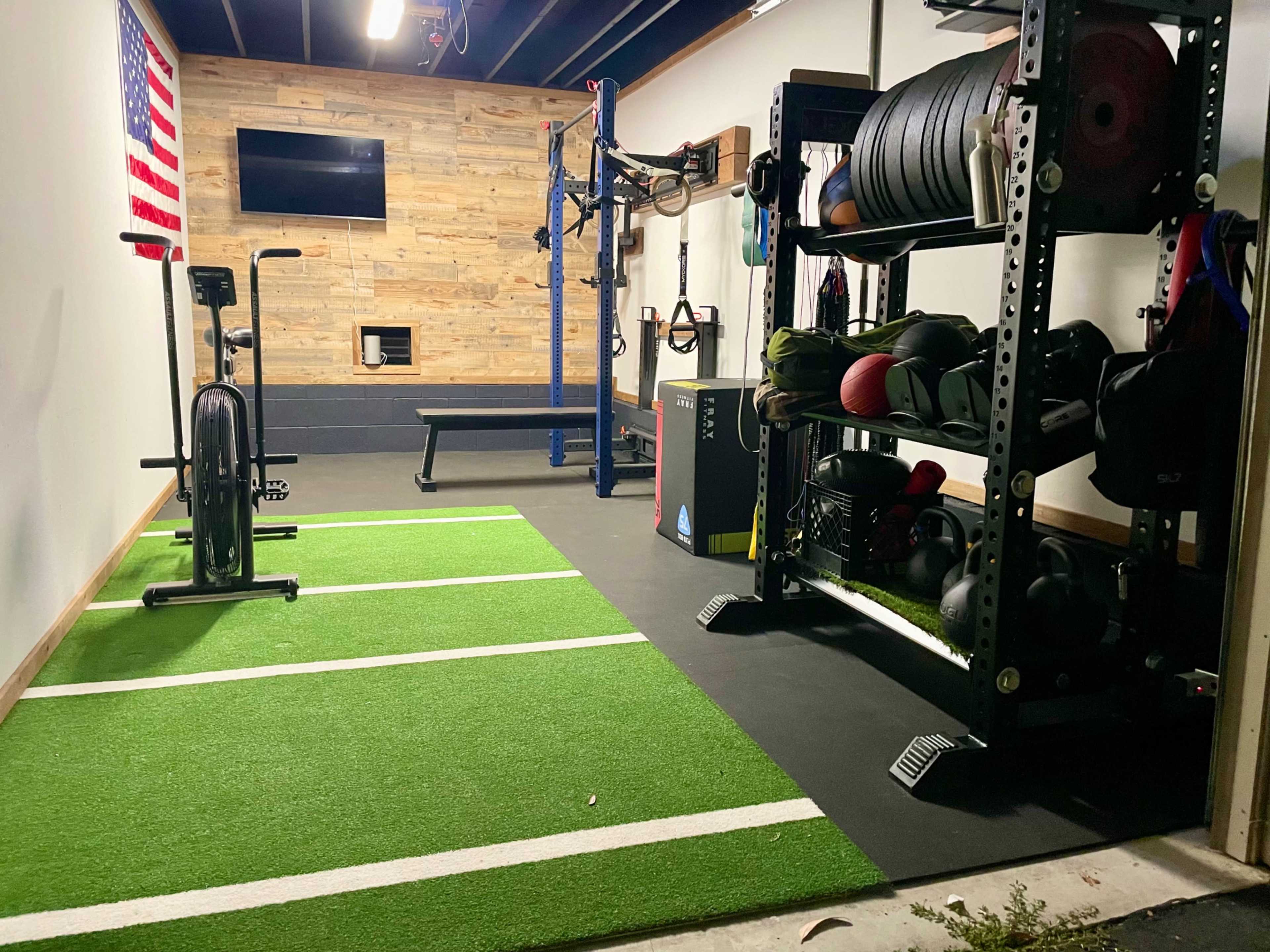 A home gym with fitness equipment including a stationary bike, weightlifting bench, and a rack of kettlebells, set against a backdrop of green turf and a wooden wall featuring an American flag.
