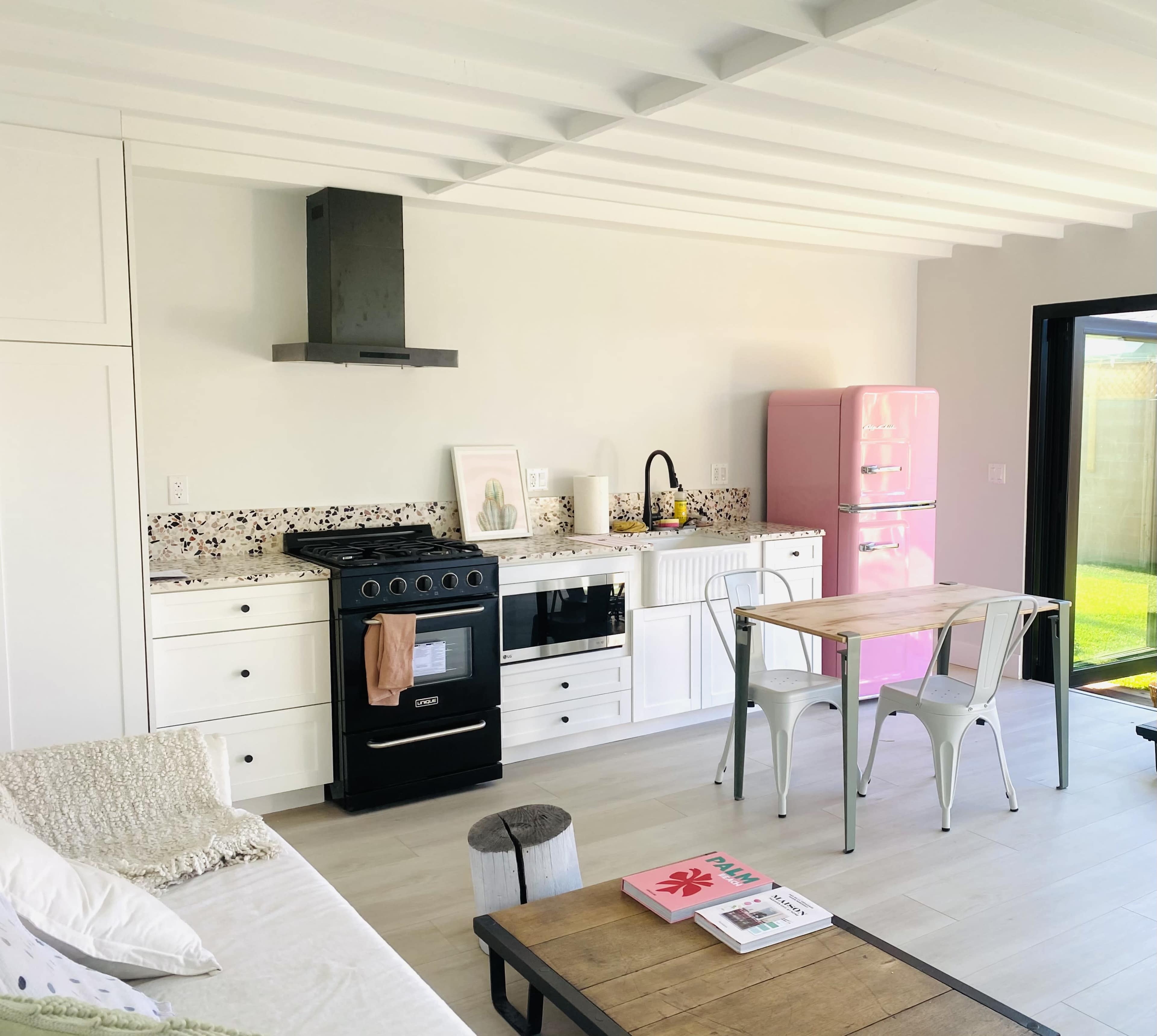 The image shows a modern kitchen and dining area featuring a black stove, white cabinetry, a pink refrigerator, a wooden dining table, and large windows allowing natural light.