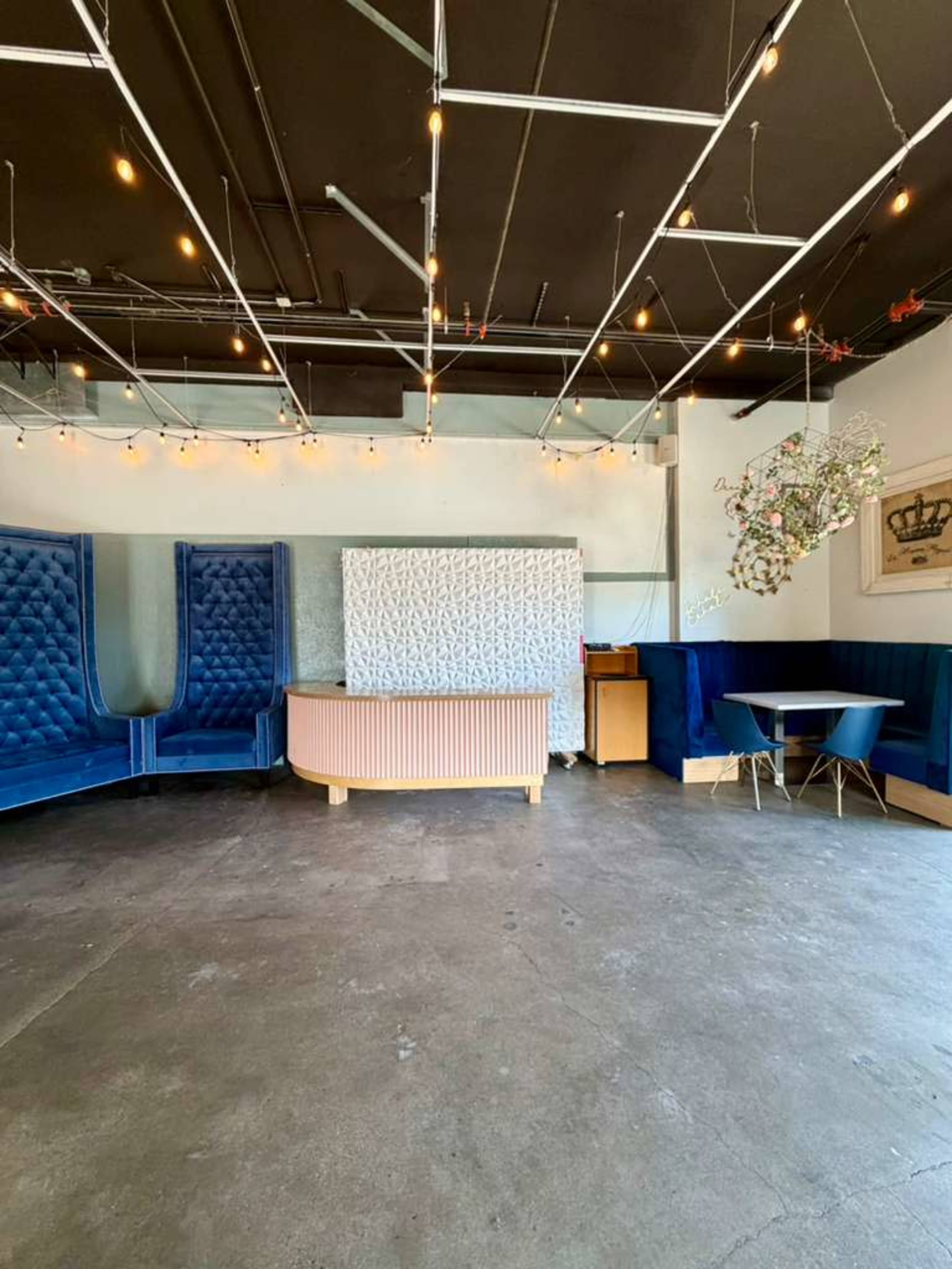 The image shows a spacious interior with blue velvet seating, a pink reception desk, and a small dining area illuminated by hanging string lights.
