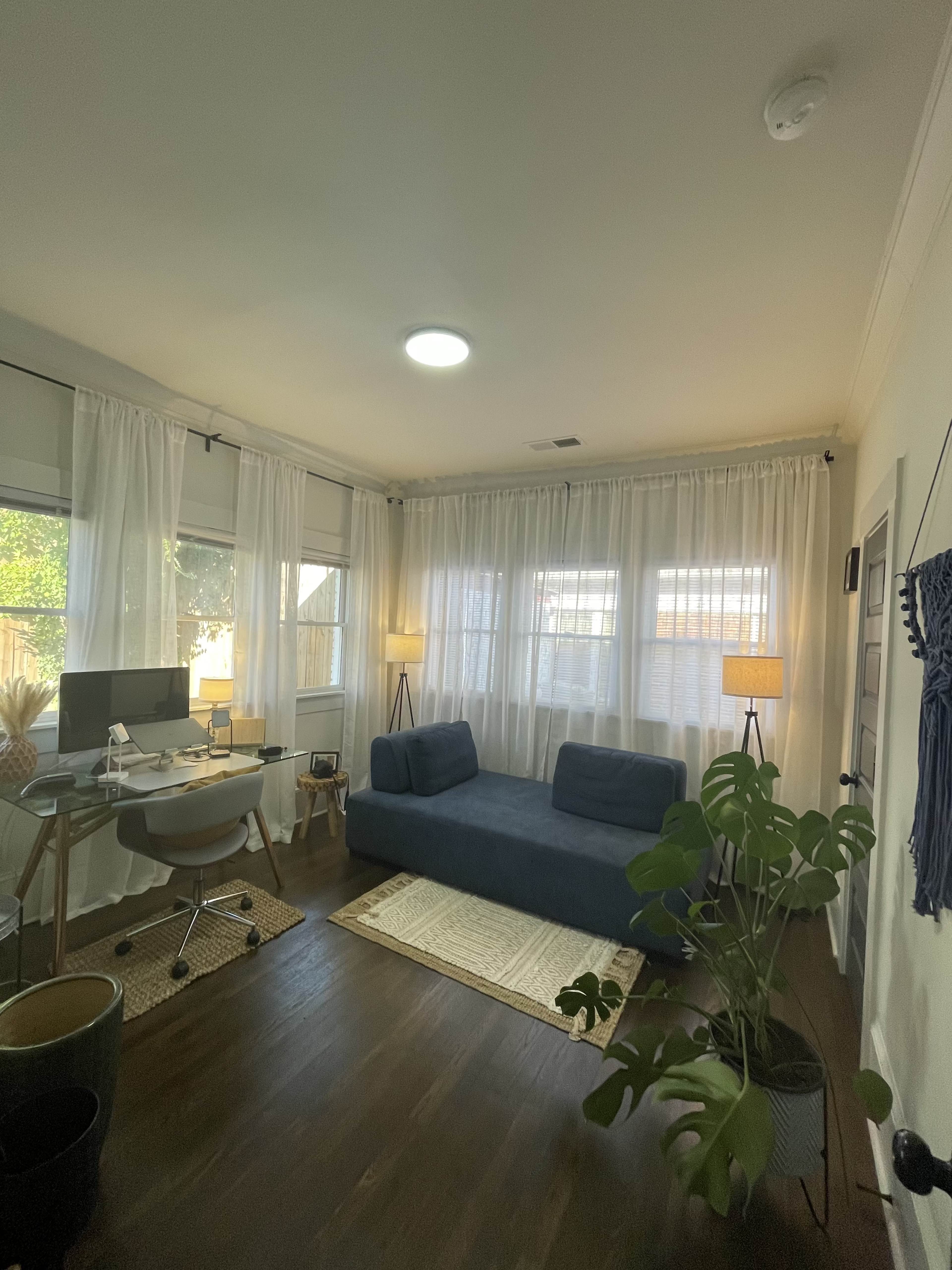The image shows a home office with a blue sofa, a desk with a computer, large windows covered with sheer curtains, and two standing lamps.