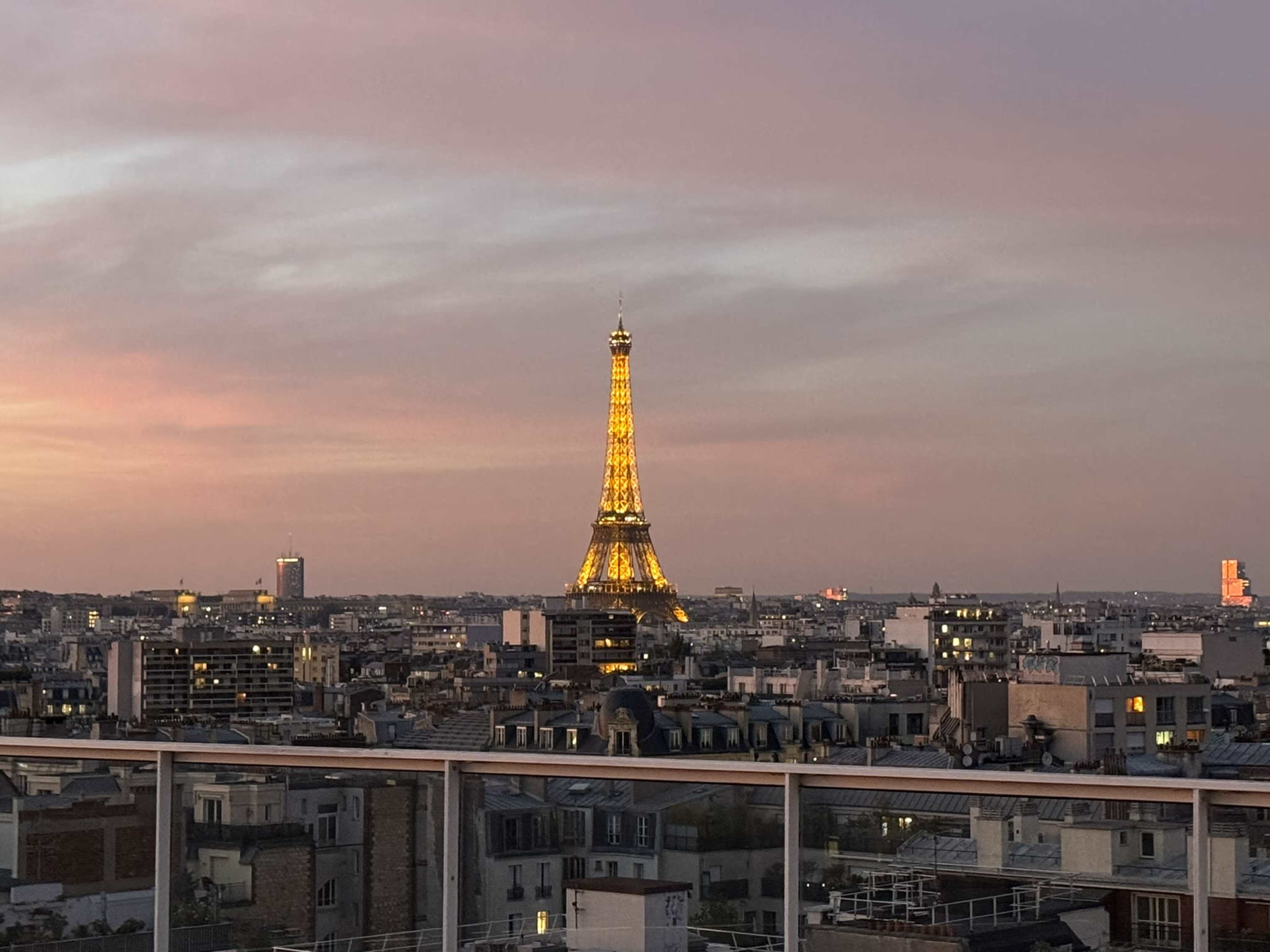 The image shows a cityscape of Paris at dusk, featuring the illuminated Eiffel Tower in the distance against a colorful sky.