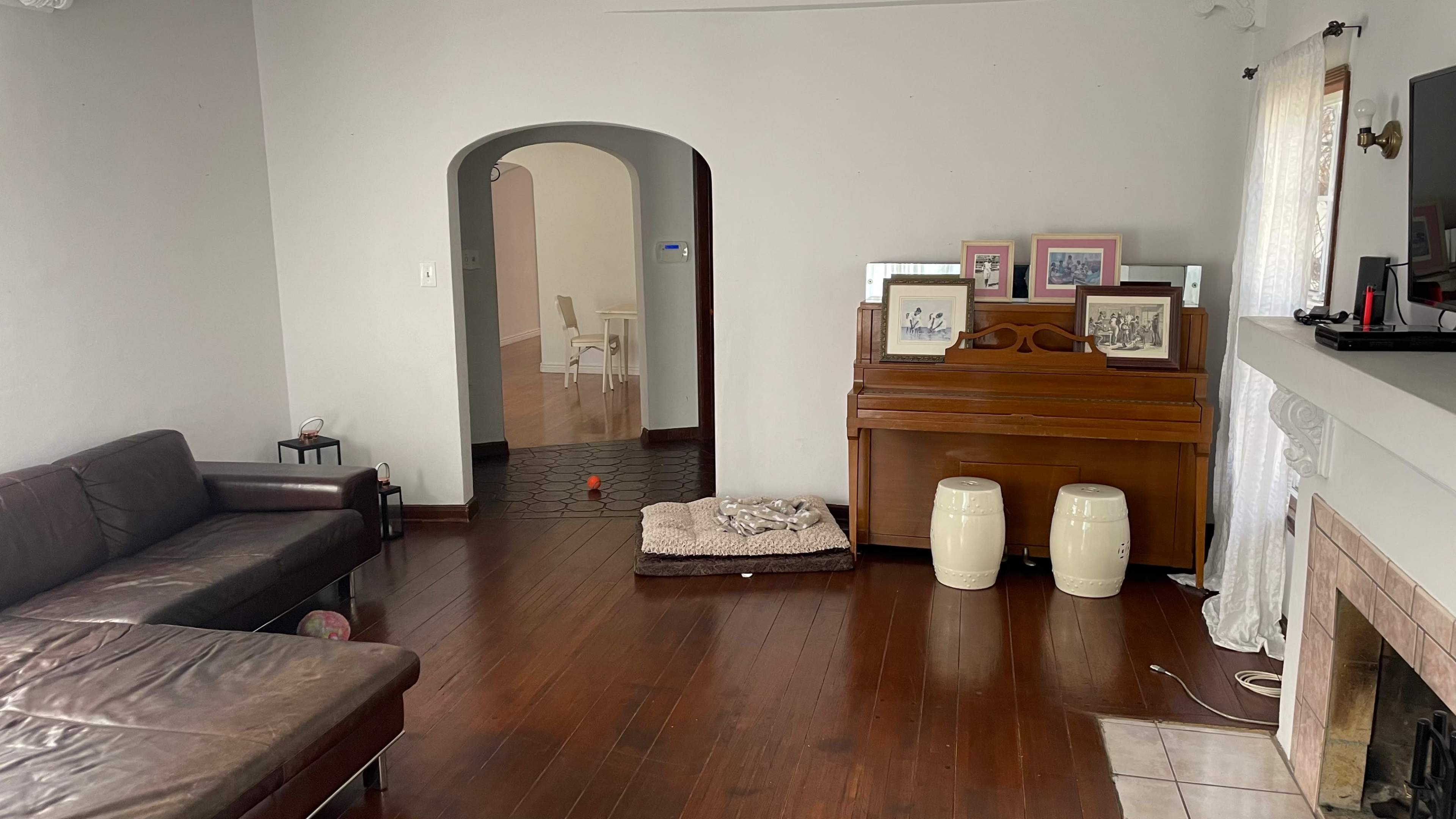 A living room with a brown leather sofa, a wooden piano with framed pictures on top, and a pet bed on the floor, leading to an arched doorway that opens to another room.