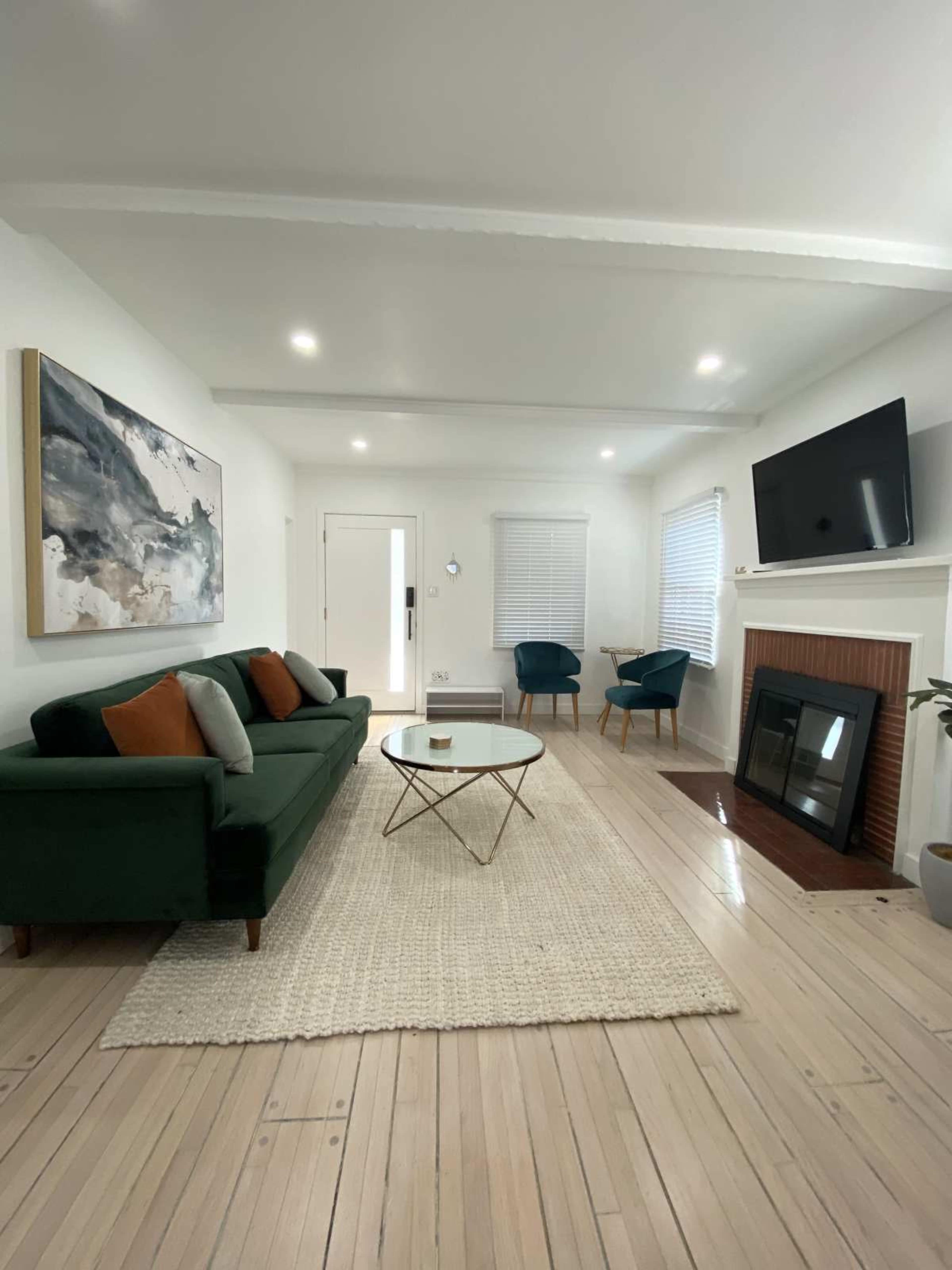 The image shows a modern living room featuring a green sofa with decorative pillows, a round coffee table, and a fireplace, with two blue accent chairs positioned nearby.