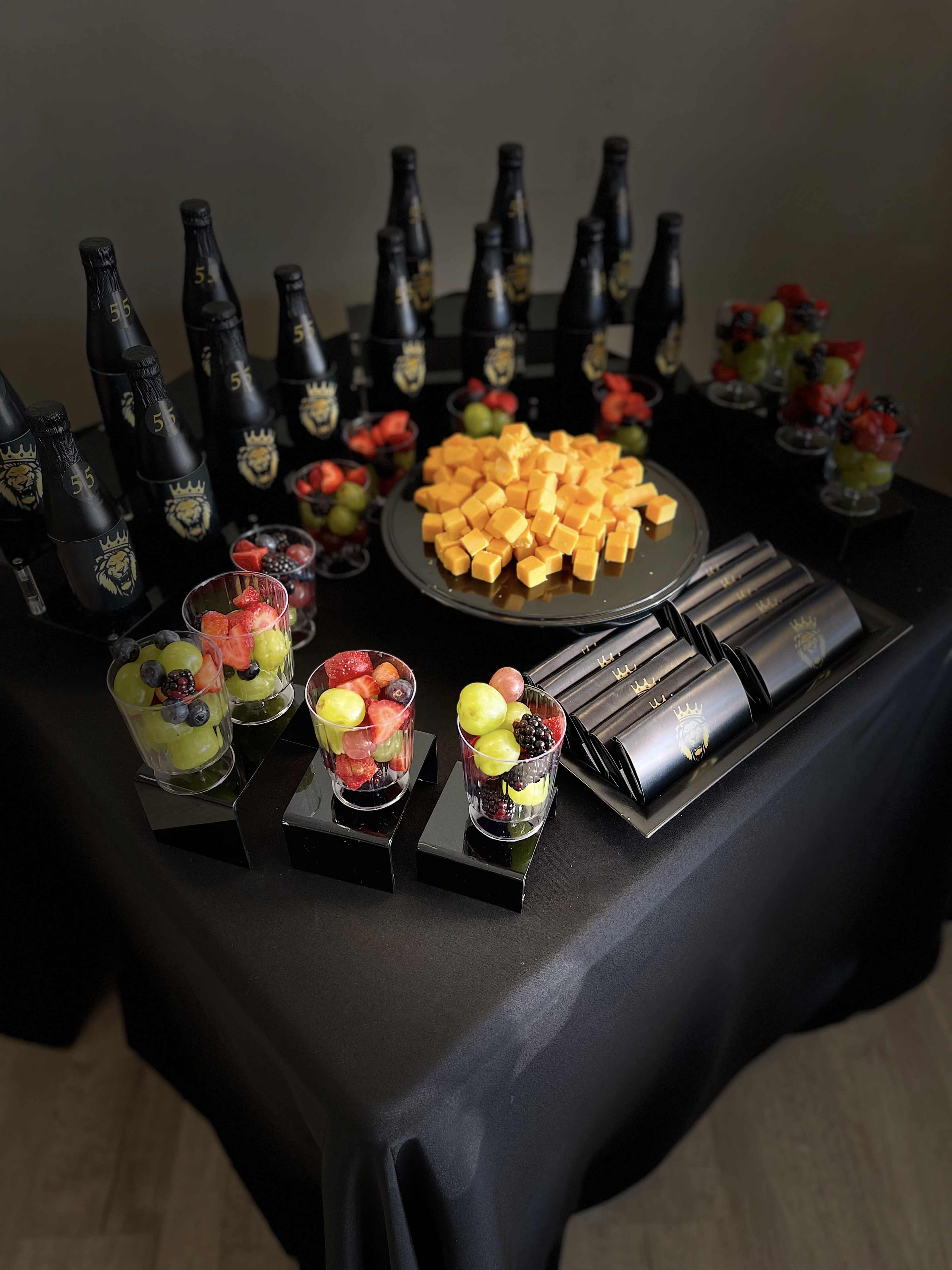 A black table is set with bottles of champagne, colorful fruit desserts in glass cups, a plate of cheese cubes, and neatly arranged black napkins.