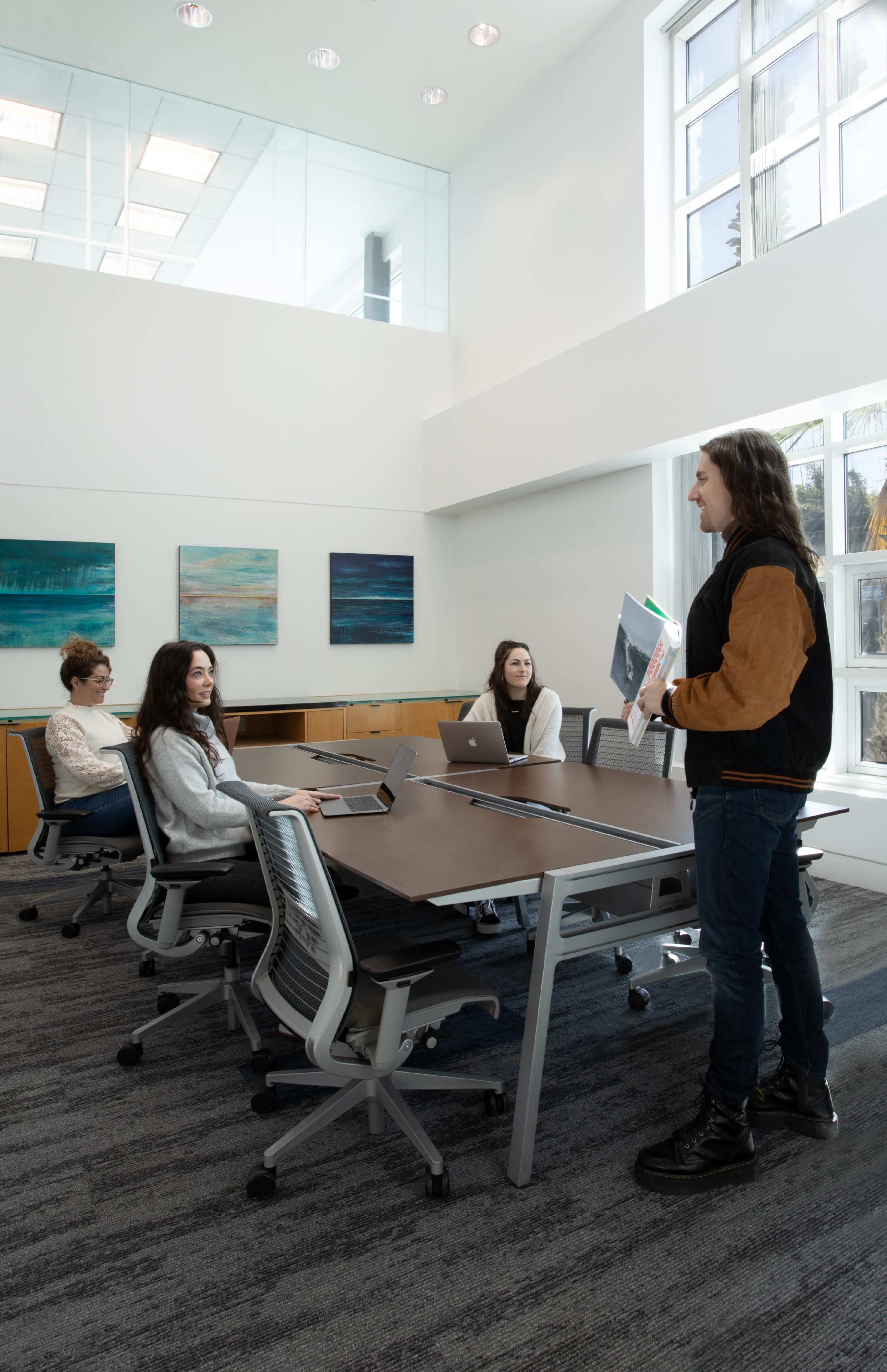 A group of four people is gathered in a modern conference room, with two individuals seated at a table and two others standing, discussing materials related to a project.