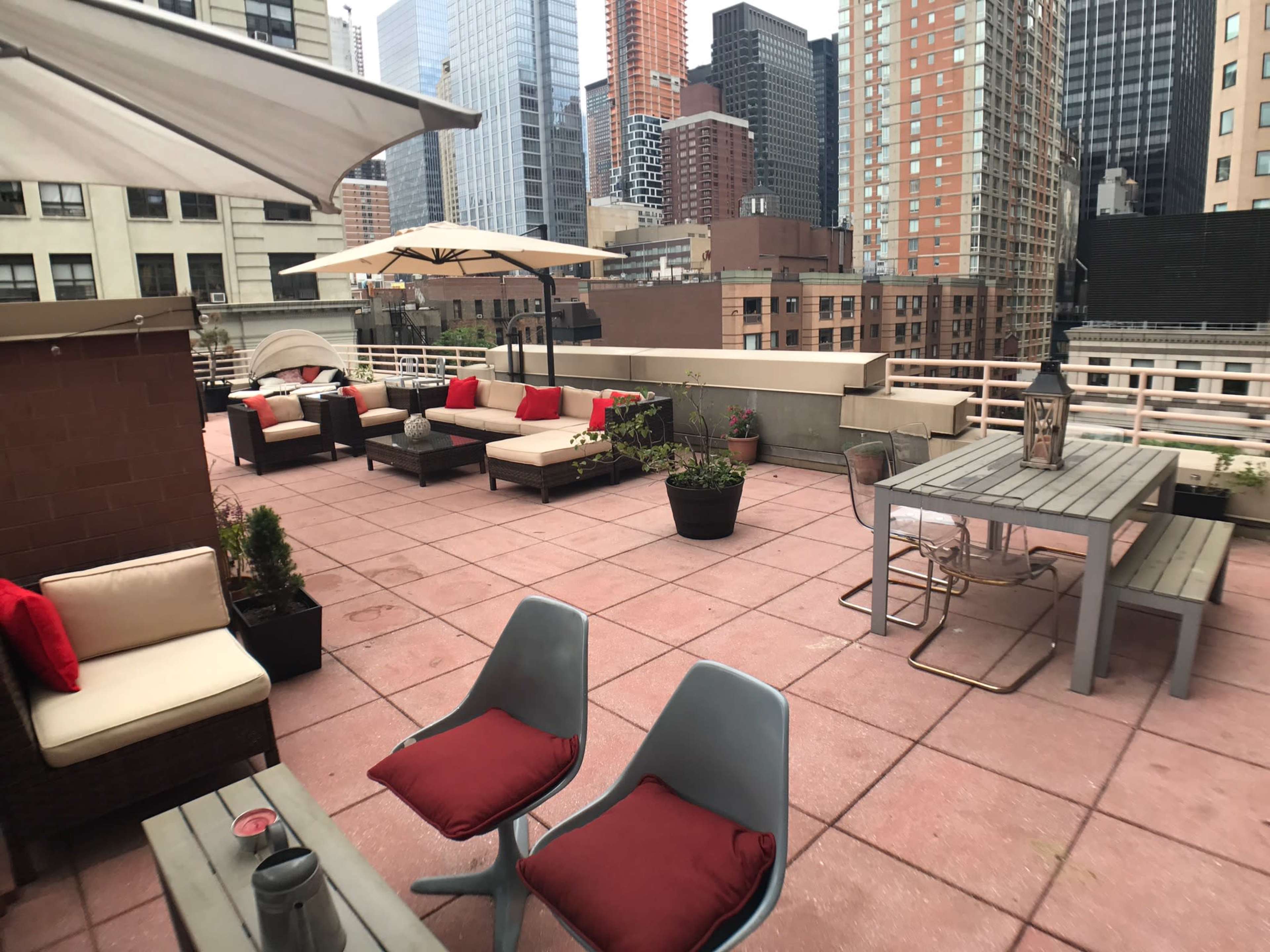 A rooftop terrace with lounge seating, a dining table, and potted plants, surrounded by tall city buildings.