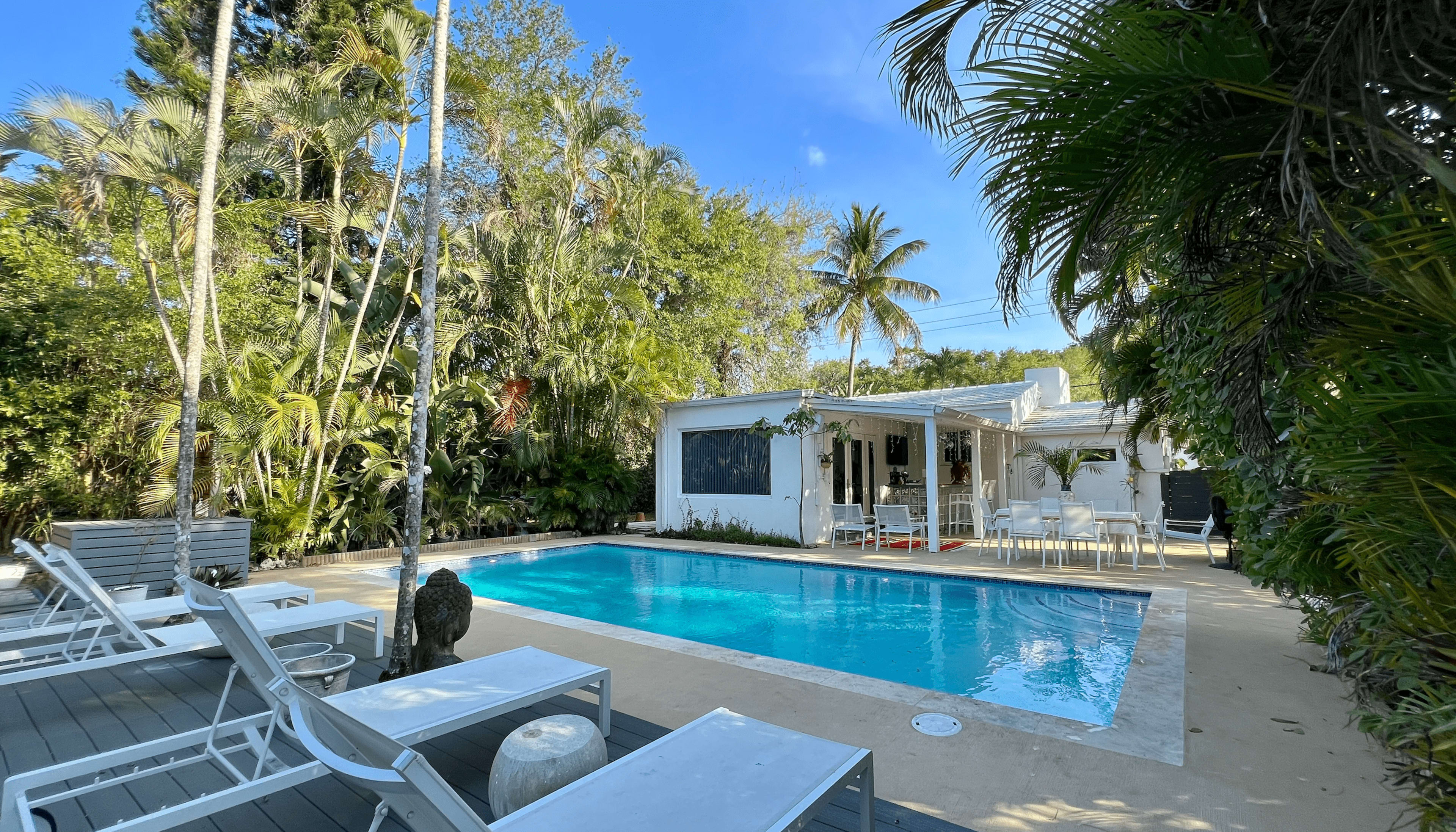 A swimming pool surrounded by lounge chairs and tropical foliage is located next to a white house with outdoor seating.