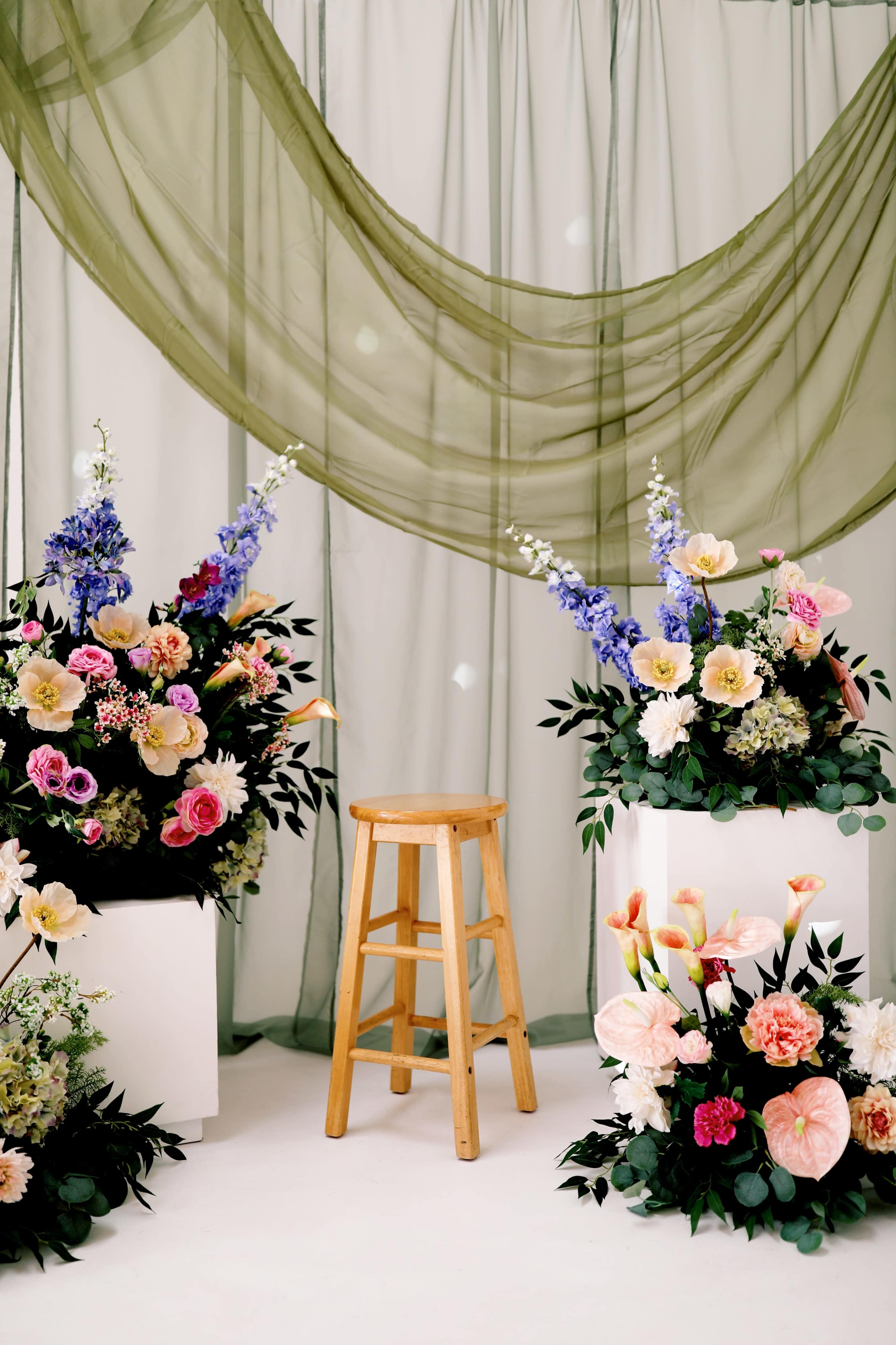 The scene features a wooden stool positioned between large floral arrangements in white containers, with flowing green drapes in the background.