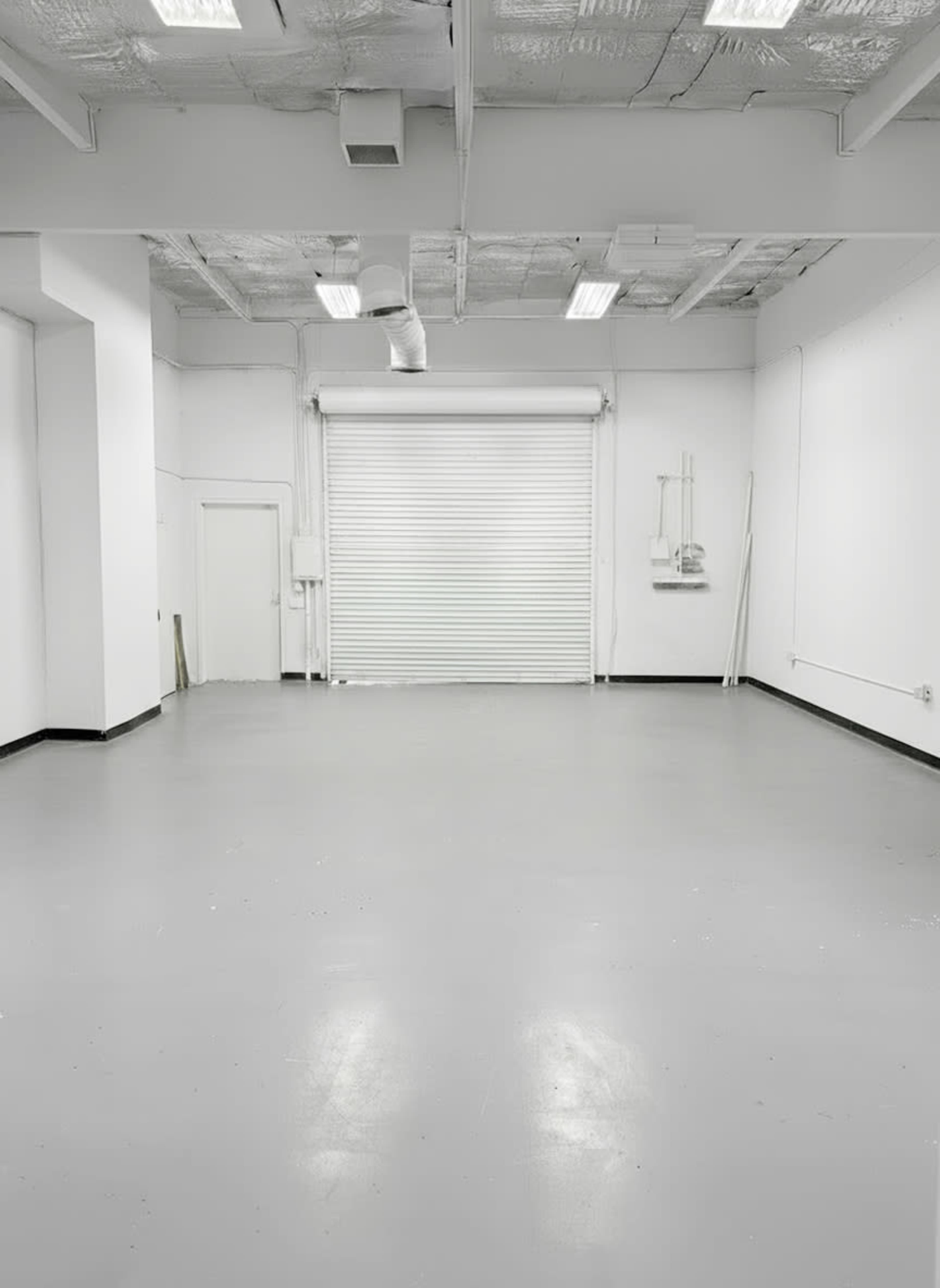 The image shows a large, empty room with a gray floor and white walls, featuring a rolled-up door at one end.