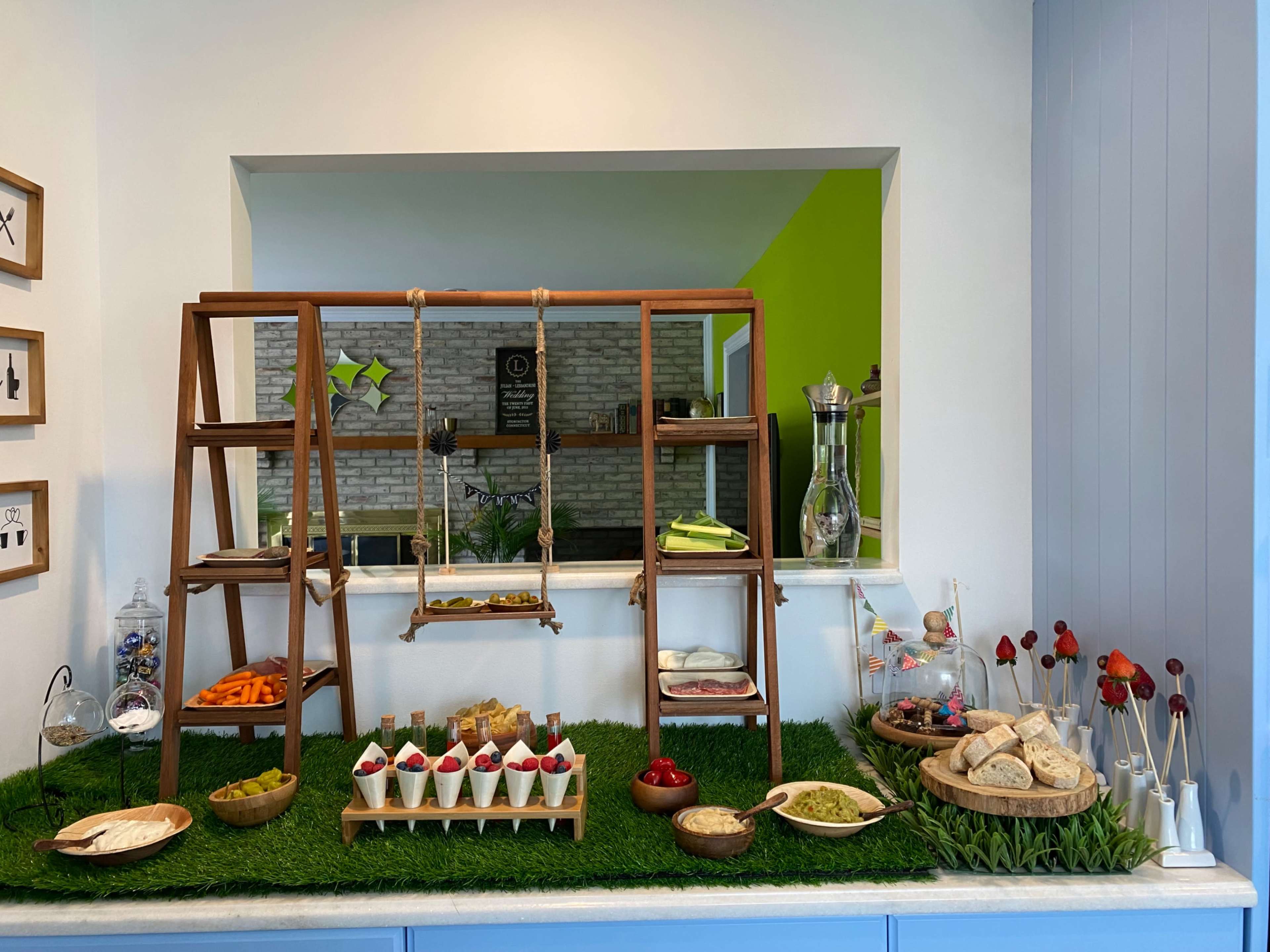 A wooden display is arranged with various dishes, including desserts, fresh vegetables, and breads, set on a grassy surface in a well-lit room.