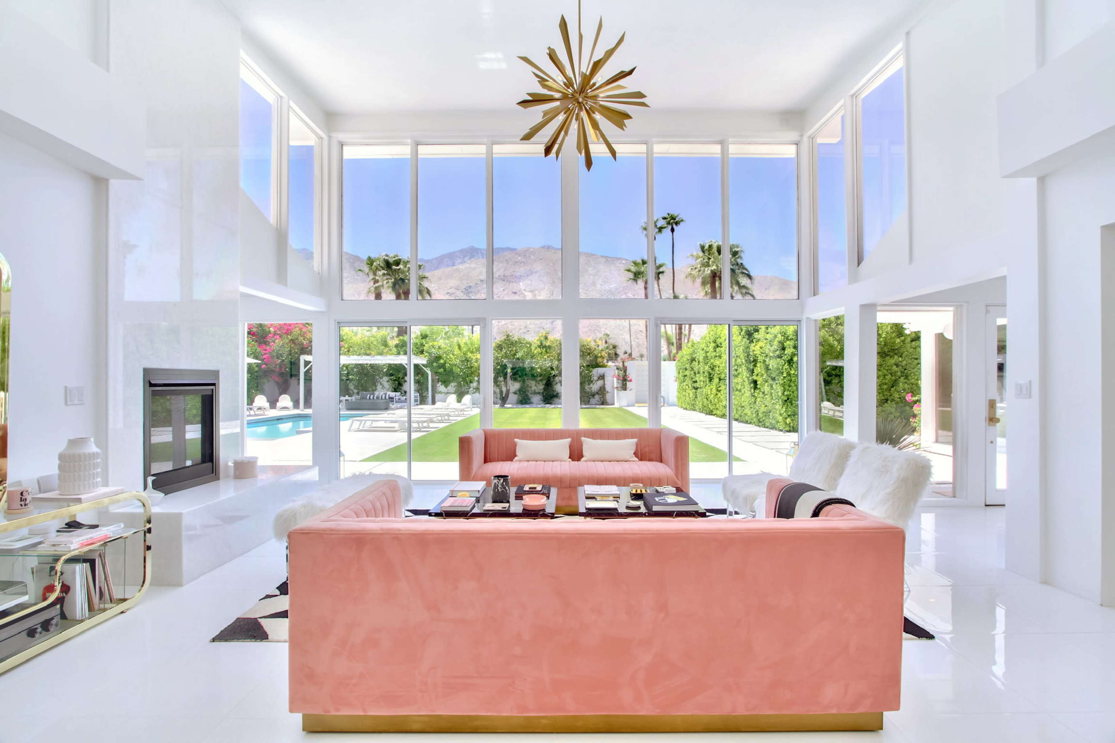 The spacious living room features large windows overlooking a pool and mountains, with pink and white sofas arranged around a coffee table.