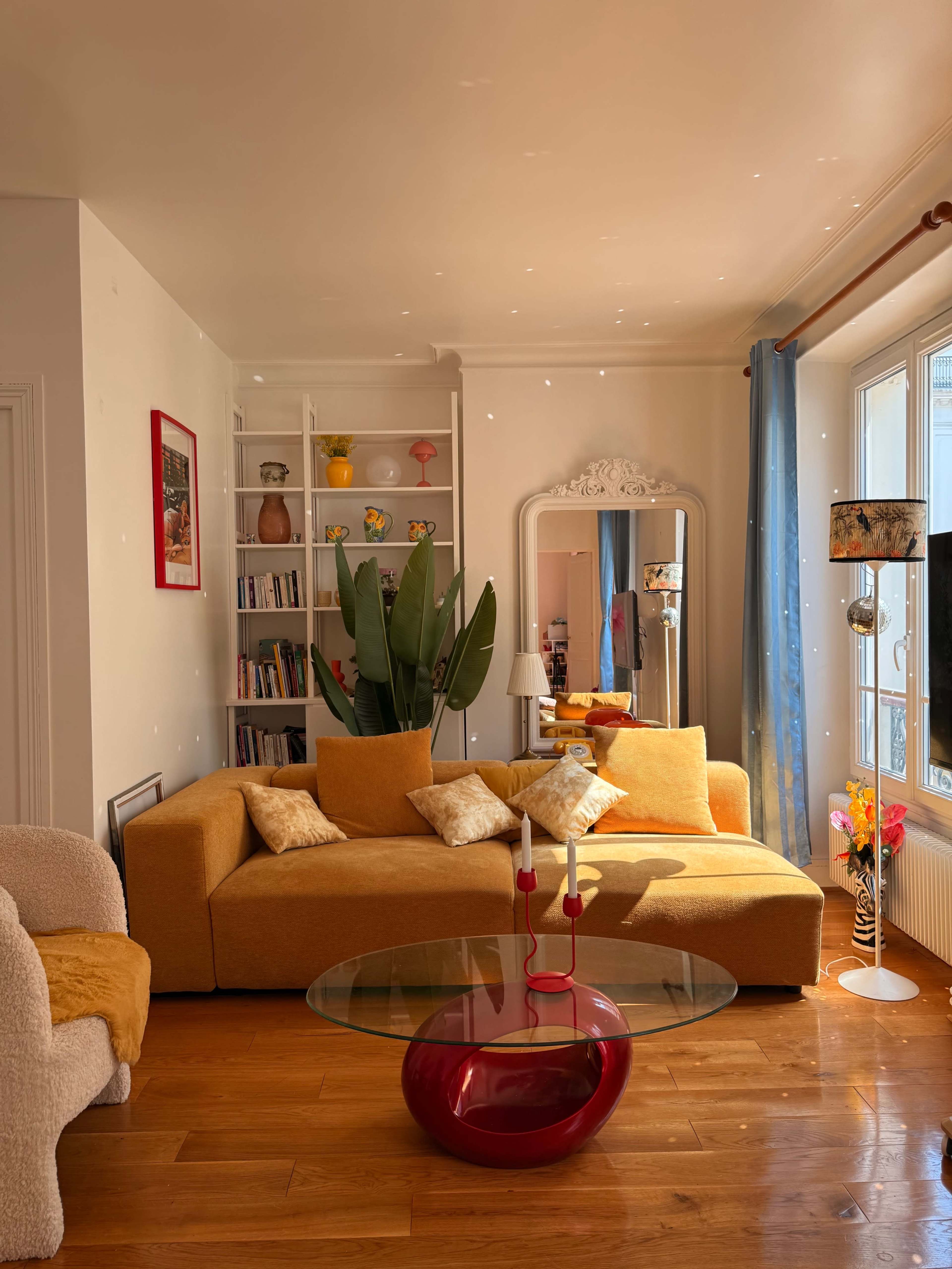 A brightly lit living room features a yellow couch, a glass coffee table, a large plant, and a decorative mirror against white walls.