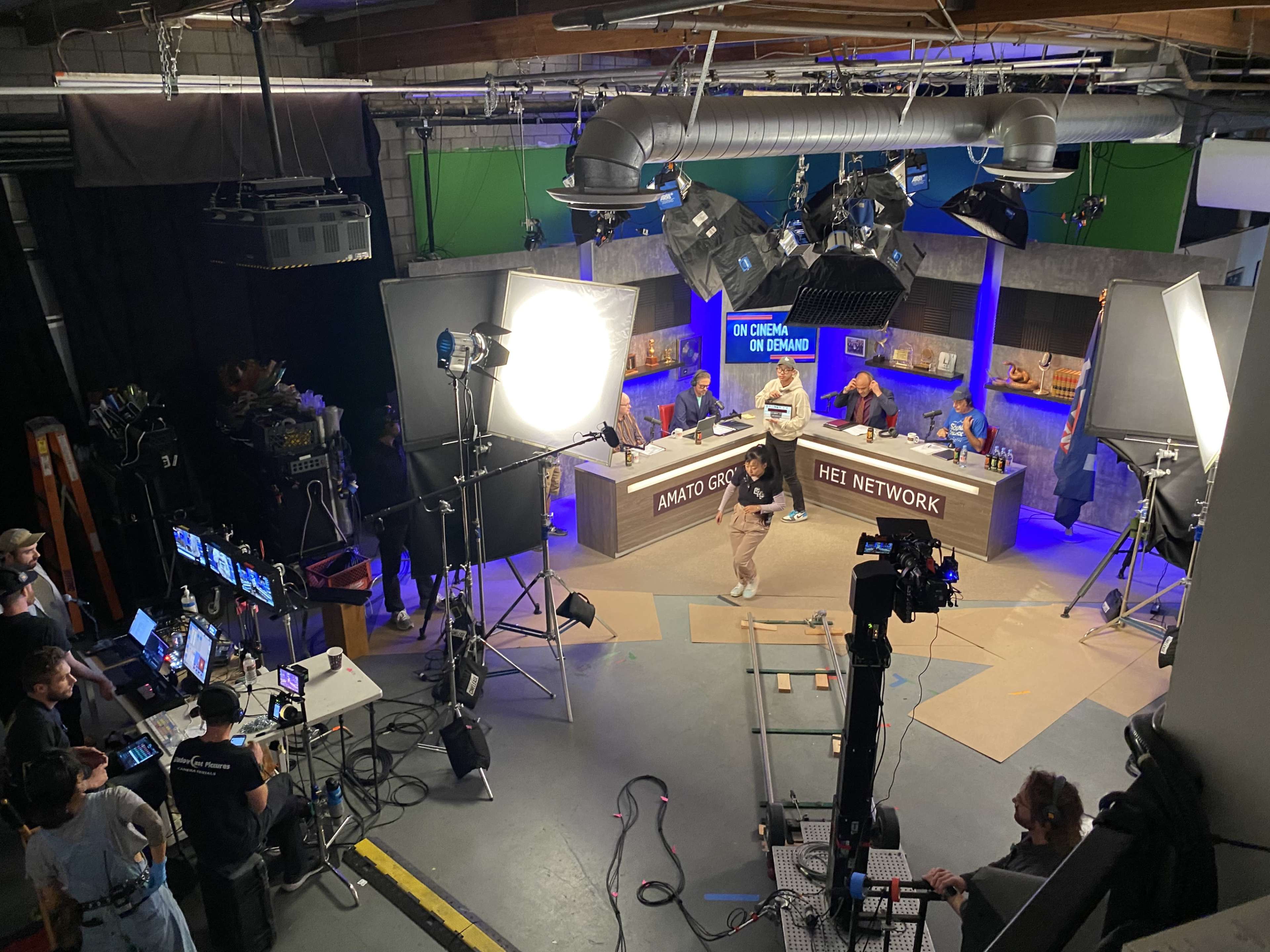 A television studio set features multiple cameras and lighting equipment, with a host walking in front of a backdrop displaying the HEI Network logo.
