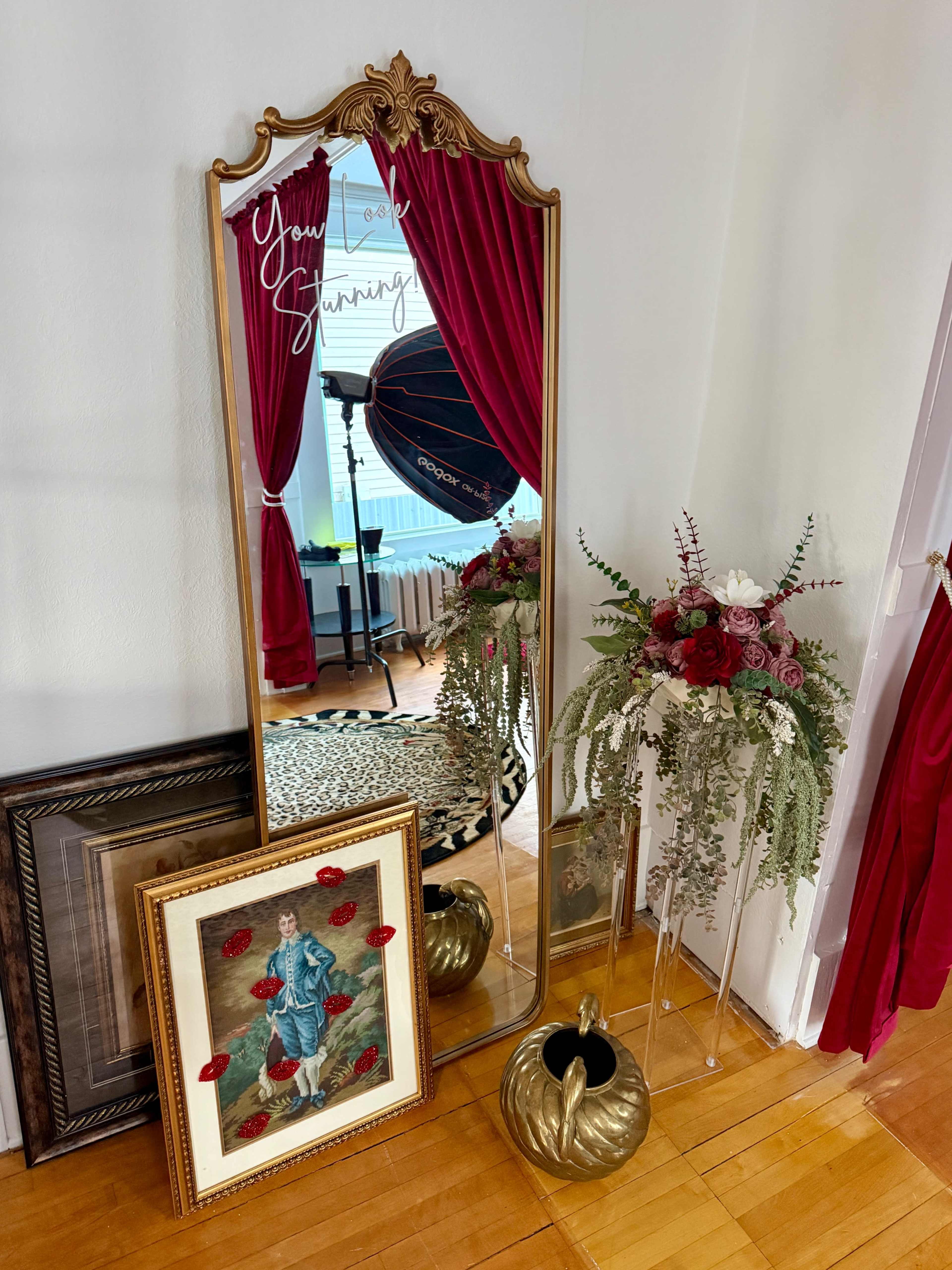 A large ornate mirror reflects a room with red curtains, a photography light, a decorative floral arrangement, and a framed painting of a woman in a blue dress.