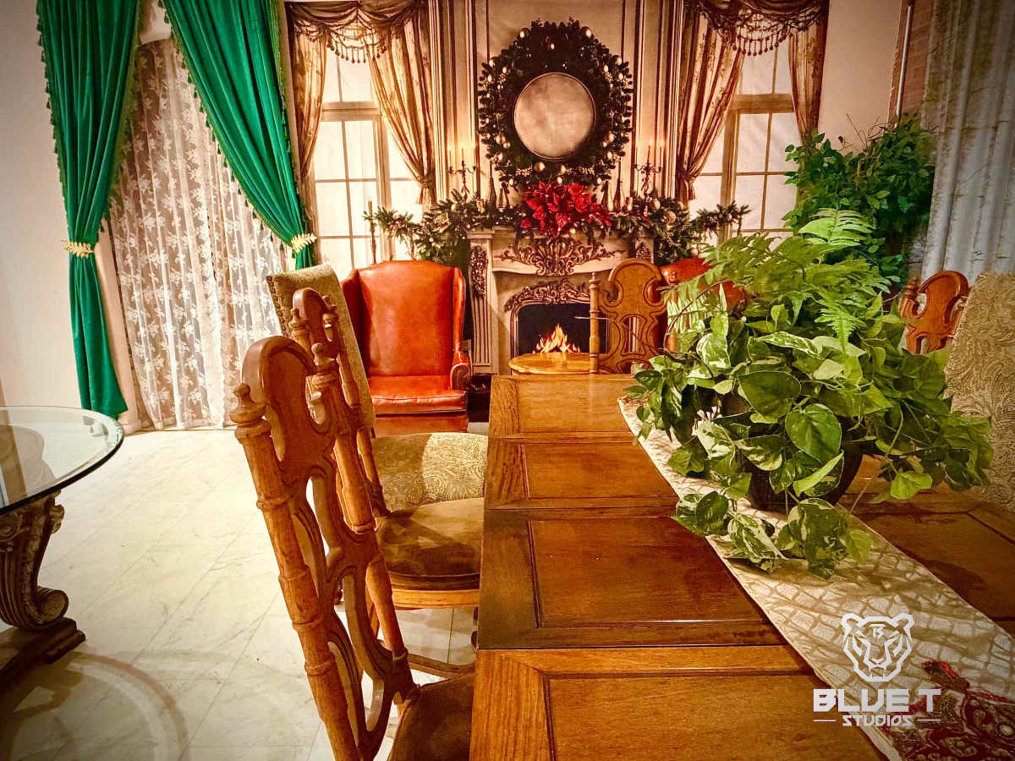 The image shows a dining area with a wooden table surrounded by ornate chairs, set against a backdrop of decorative curtains and a fireplace.