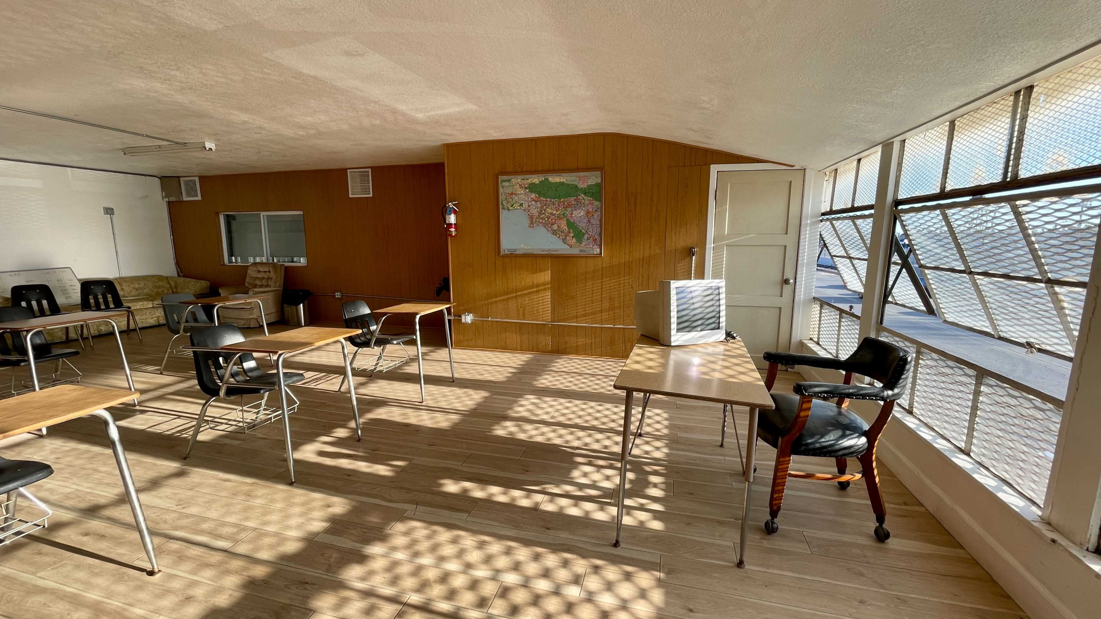 A sparsely furnished classroom with several wooden desks, a map on the wall, and large windows that allow sunlight to cast shadows on the floor.