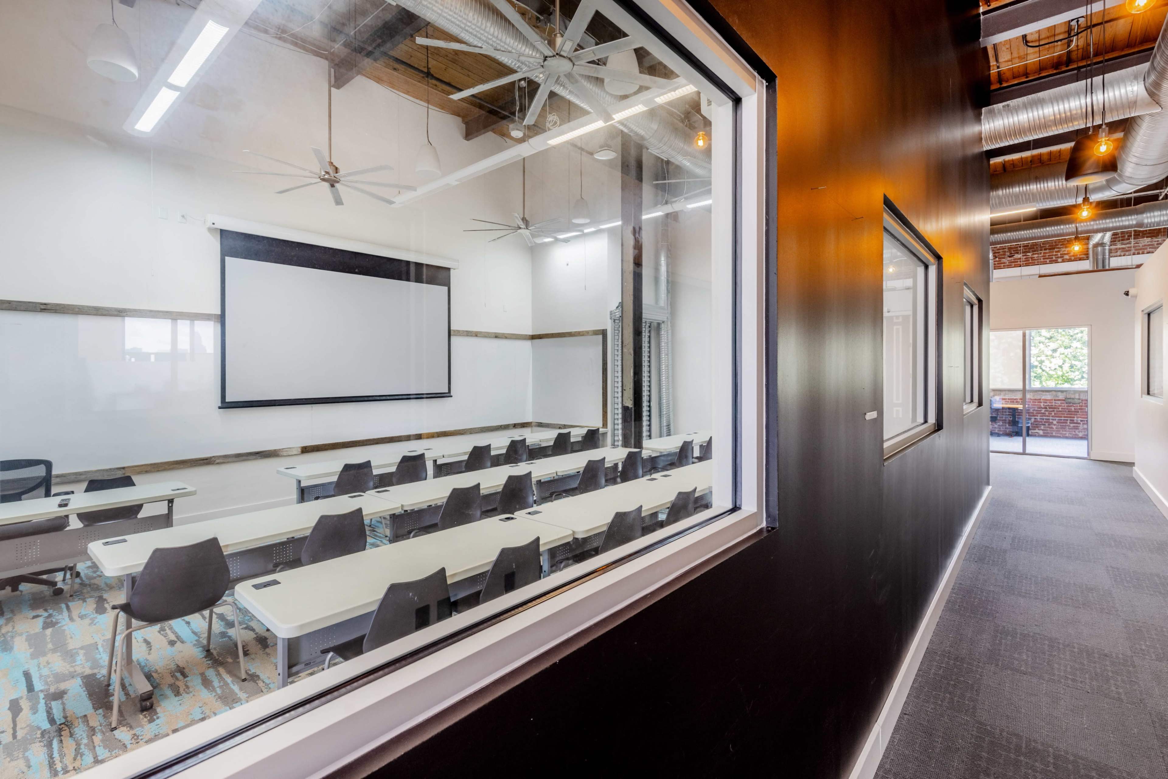 A hallway leads to a classroom featuring rows of tables and chairs, with a projector screen and large windows.