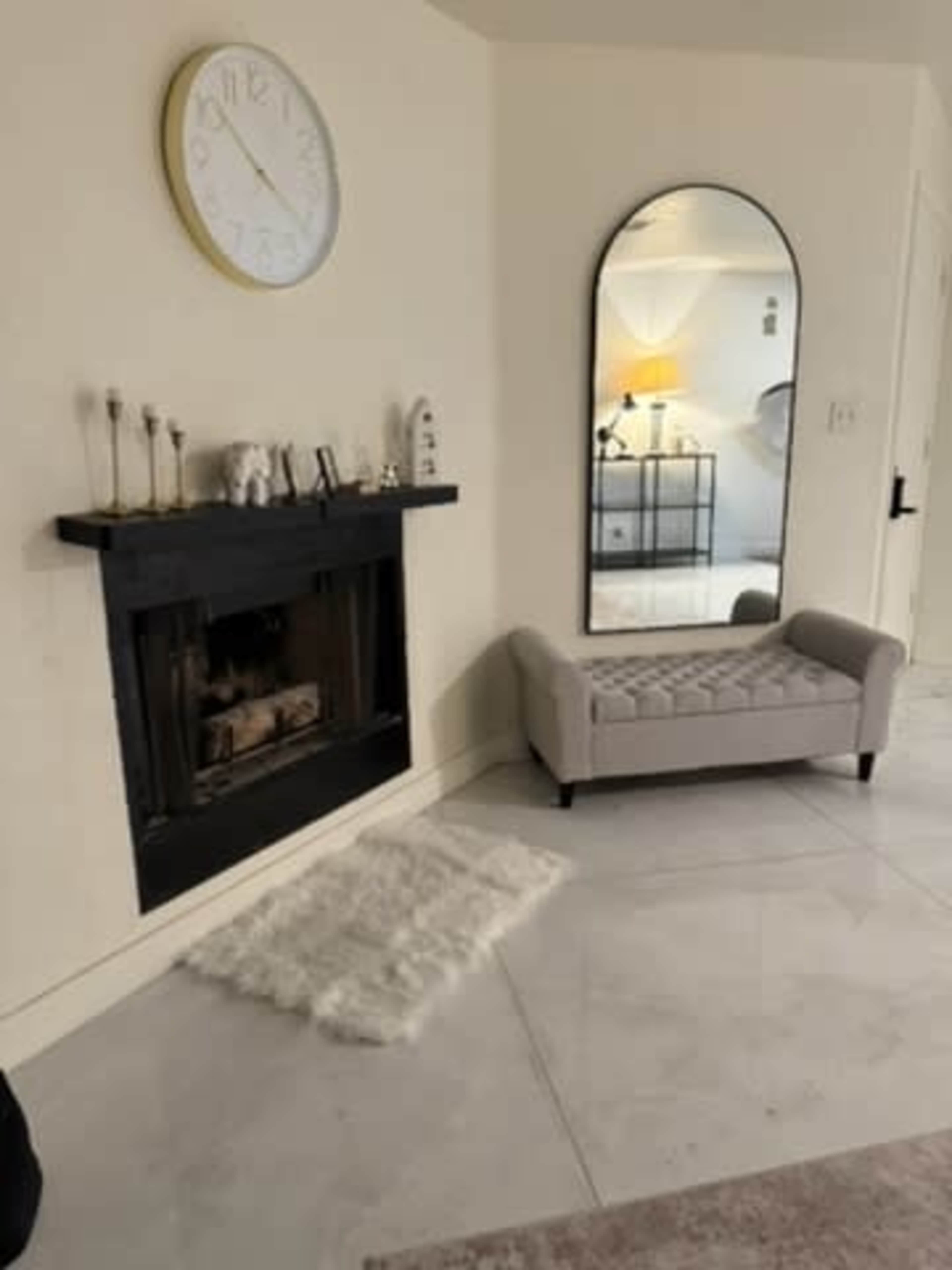 The image shows a modern living room featuring a wall-mounted clock, a black fireplace with decorative items, a tufted seating area, and a large mirror reflecting part of the space.