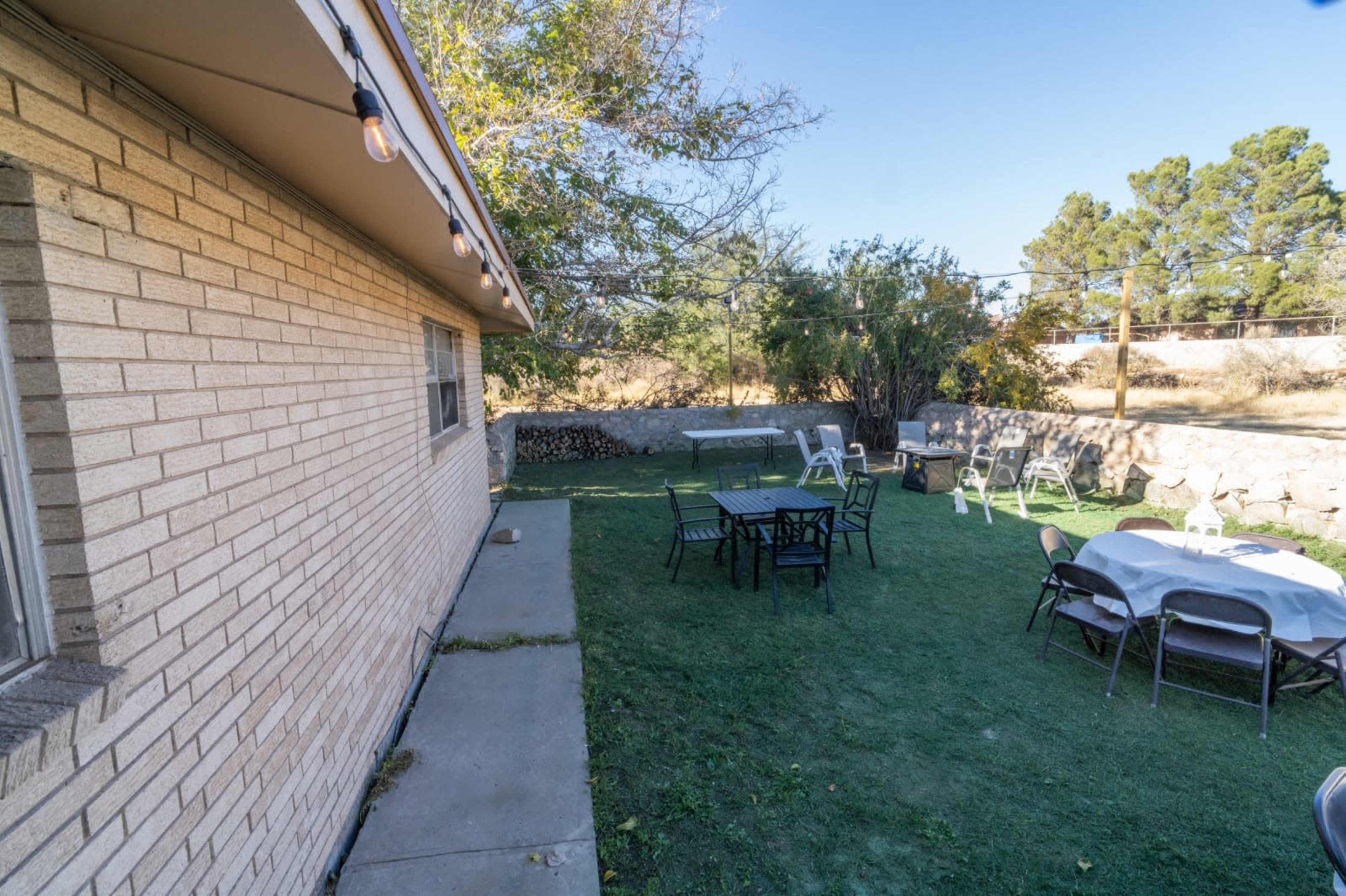 The image shows a backyard area with several tables and chairs set up on a grassy lawn next to a brick wall.
