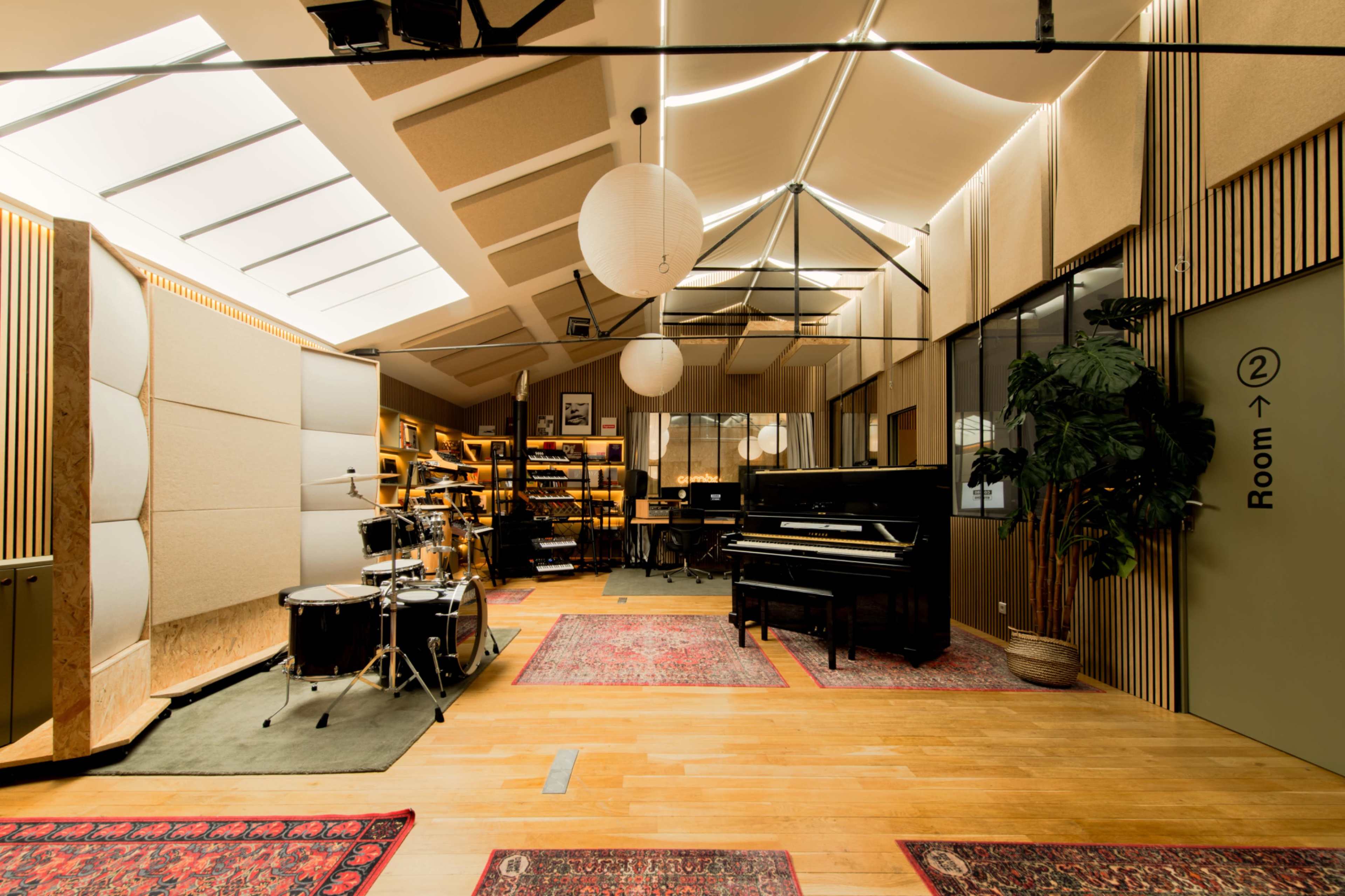The interior of a music studio features a grand piano, drums, and shelves filled with equipment, all set against a backdrop of large windows and modern decor.