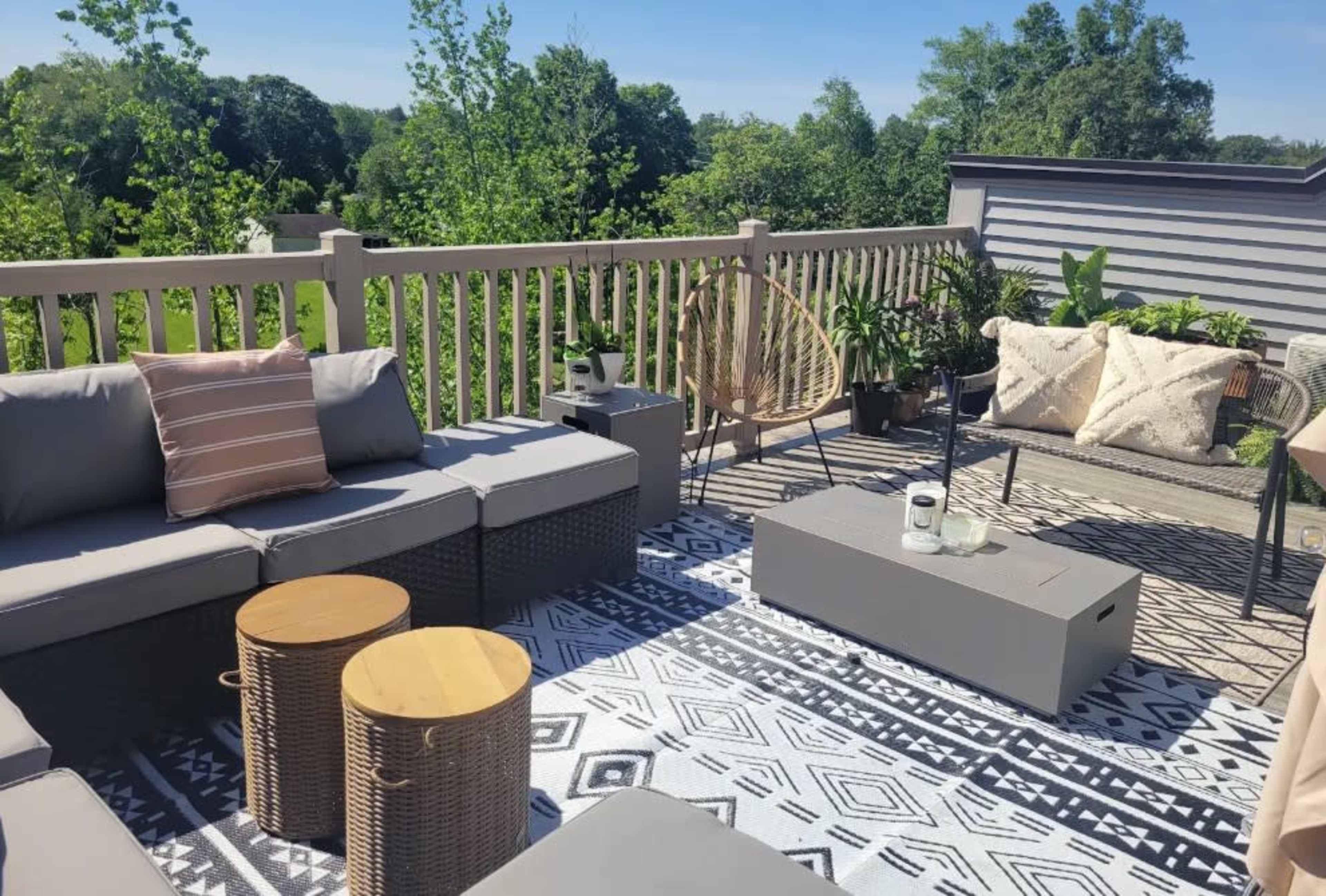 The image shows a rooftop patio furnished with a sectional sofa, wooden side tables, and decorative plants, all set on a patterned rug with a view of greenery in the background.