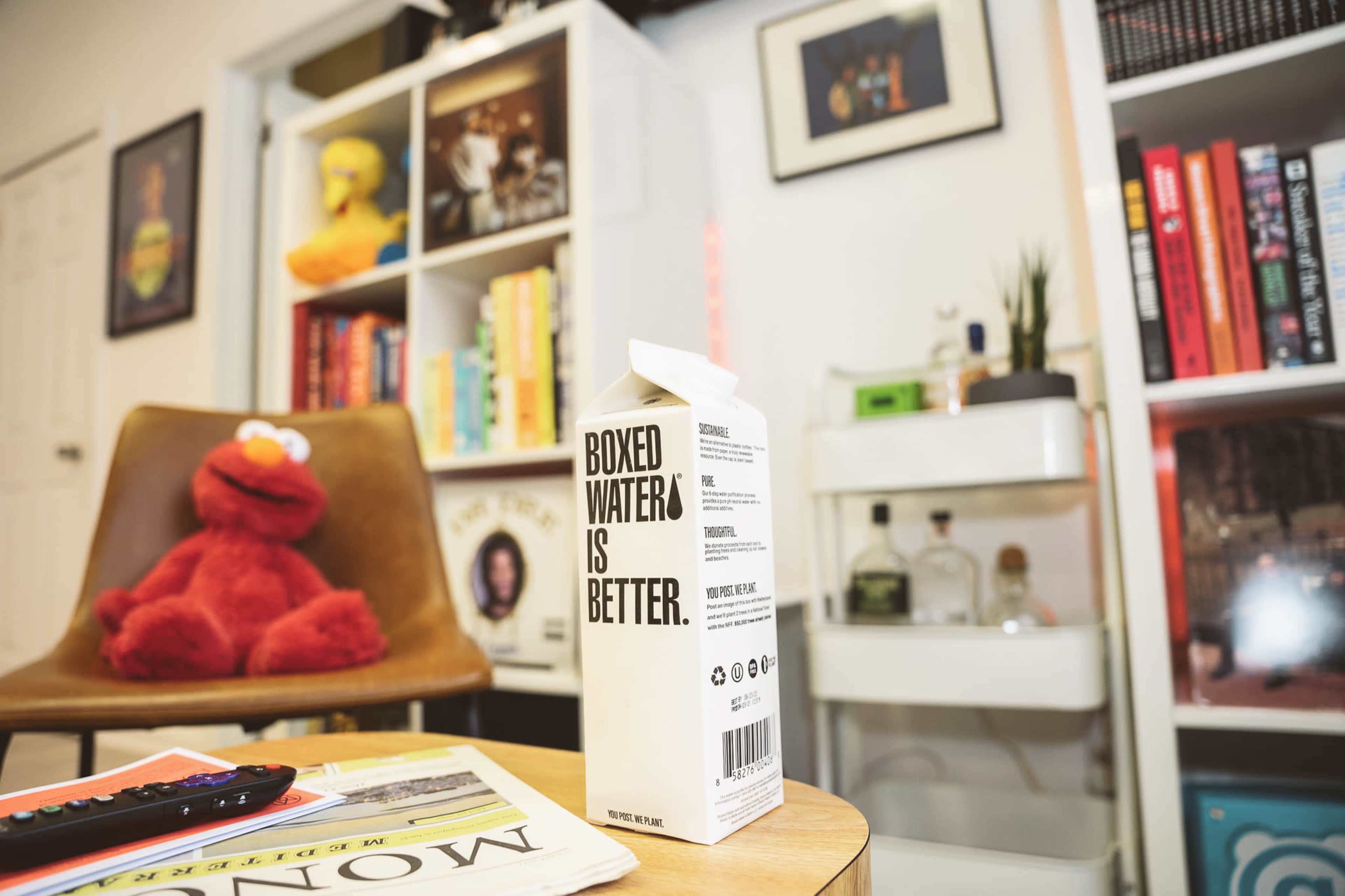 A carton labeled "BOXED WATER IS BETTER" sits on a coffee table next to a remote control, with a plush red character in the background and bookshelves filled with colorful books.
