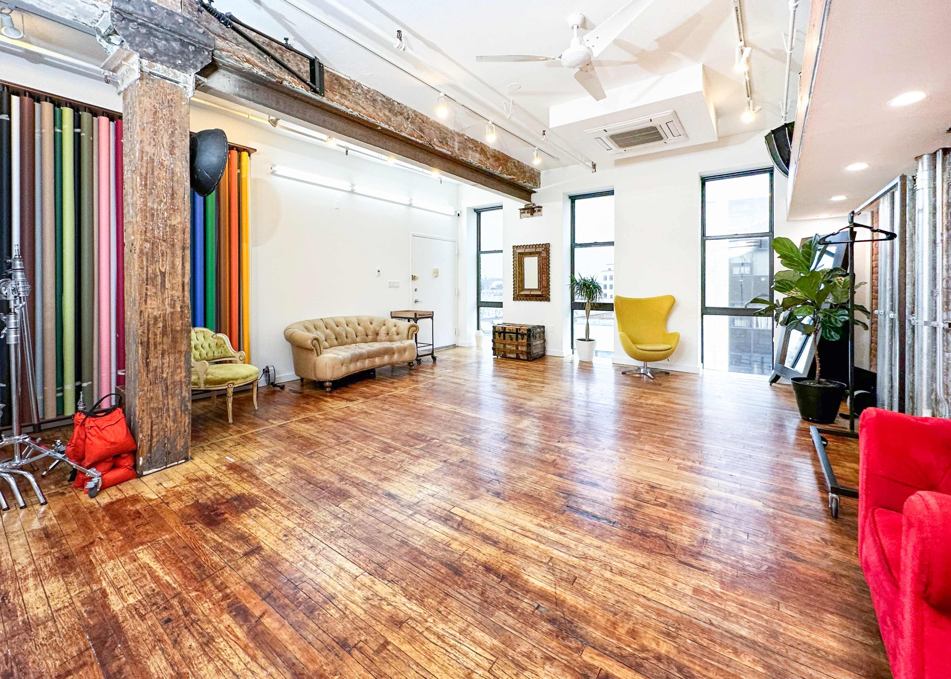 A spacious, well-lit room featuring wooden floors, exposed beams, various seating options, and colorful vertical backdrops along one wall.